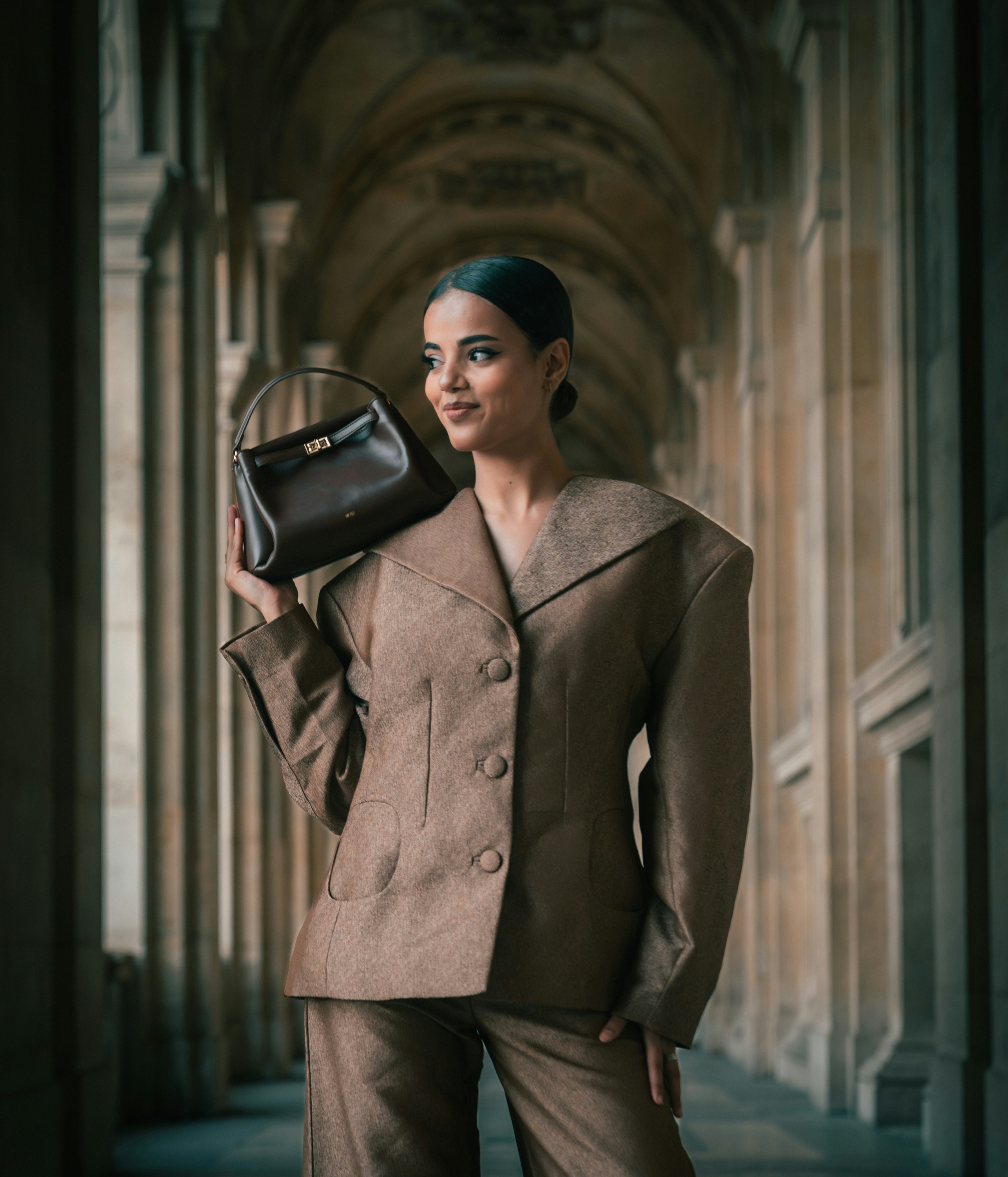 A woman in a brown suit holding a dark handbag in a stone corridor