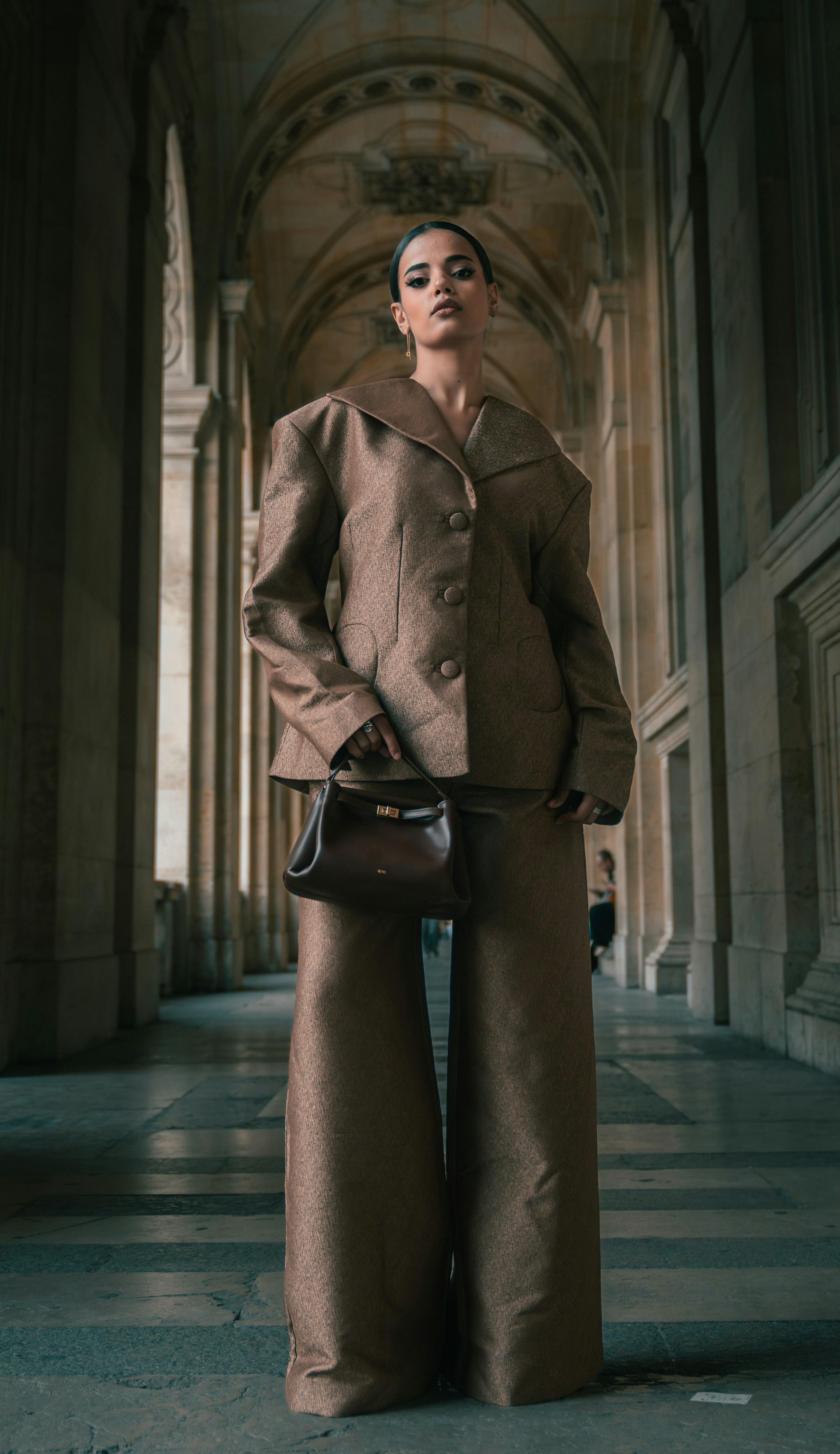 A woman in a brown suit holding a dark handbag in a stone corridor