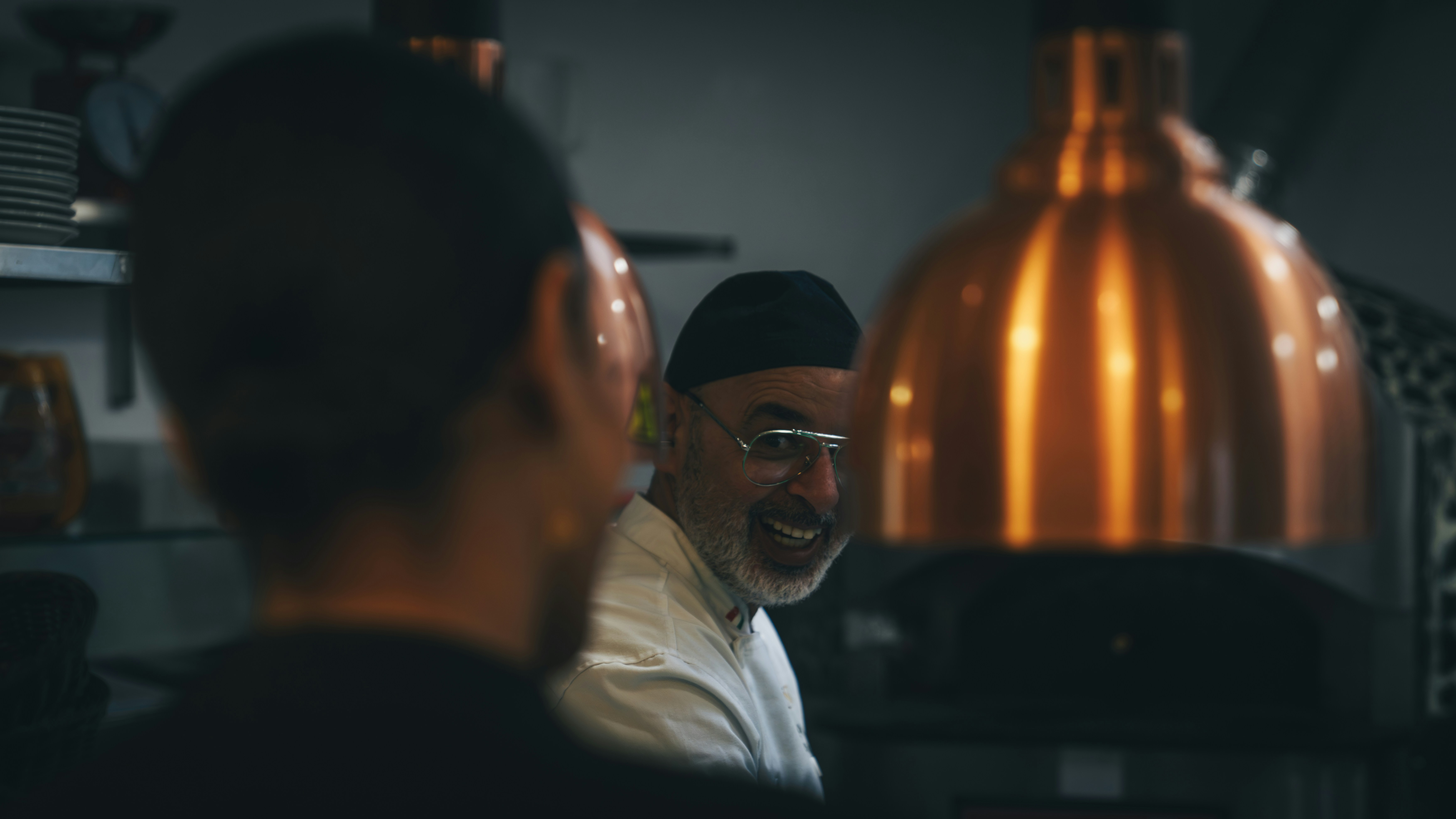 A smiling chef in a white coat and black cap in a kitchen