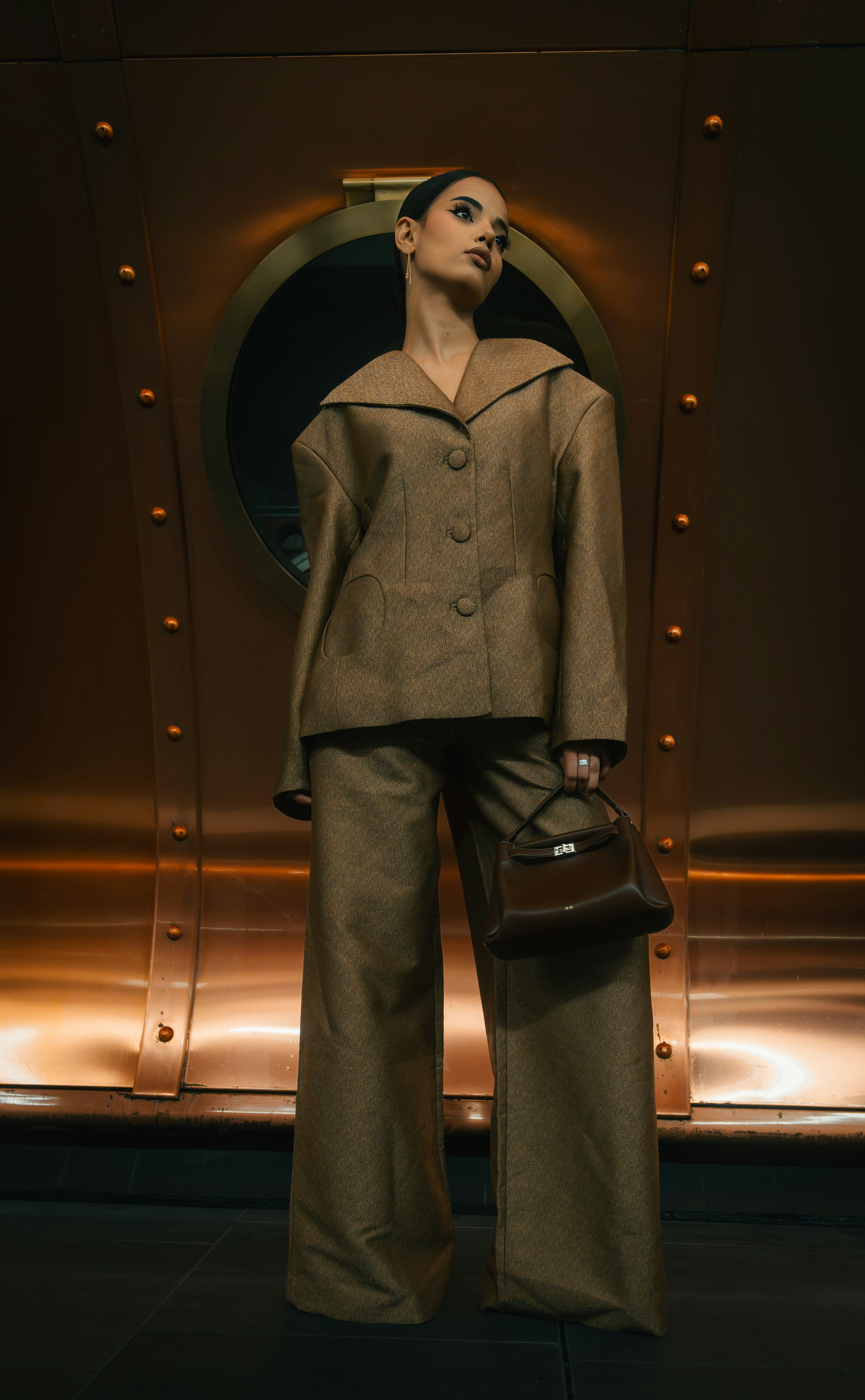 A woman in a brown suit holding a dark handbag against a copper wall