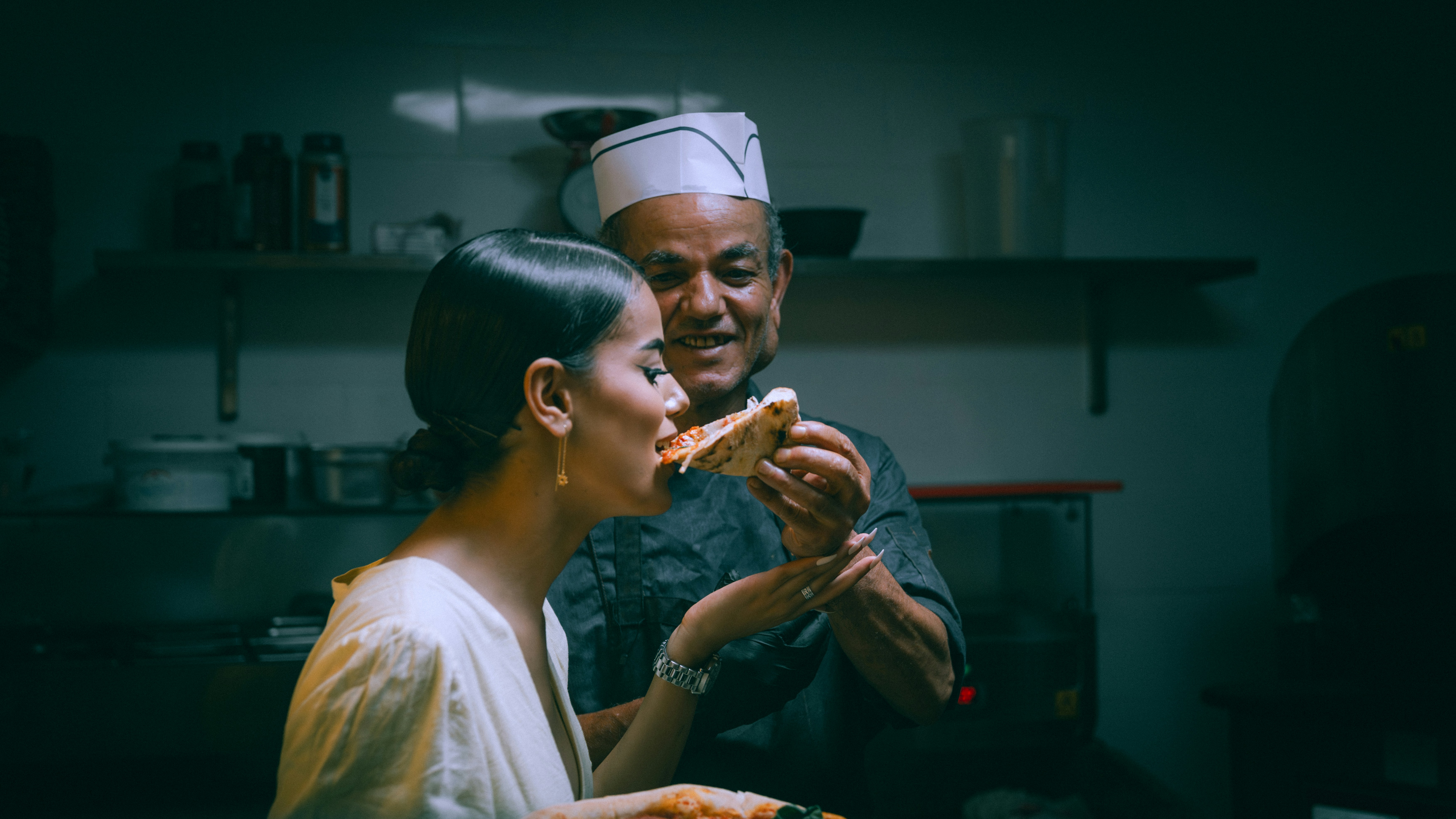 A chef in a white hat feeding a woman a slice of pizza
