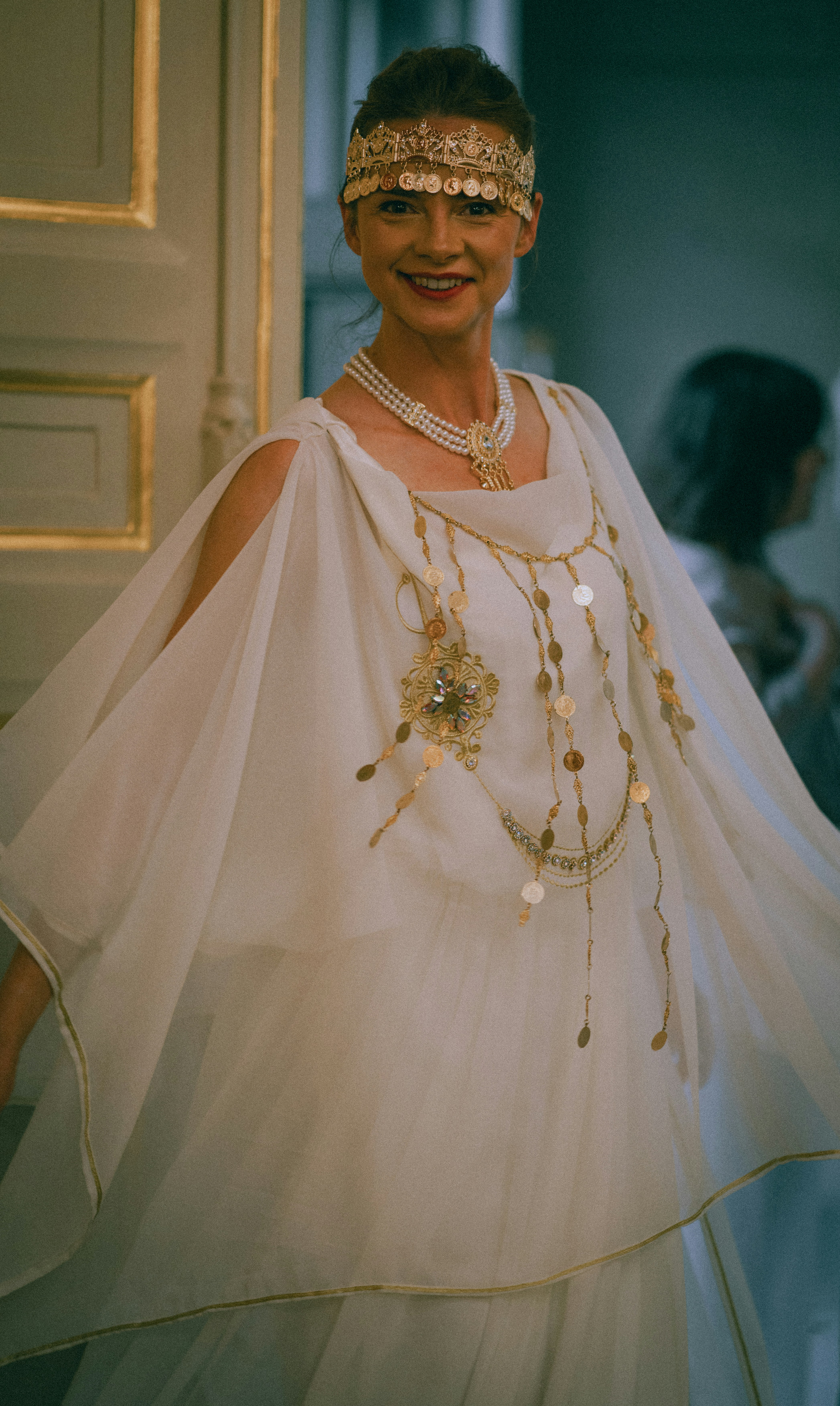 A woman wearing a white draped dress and gold coin headpiece and jewelry