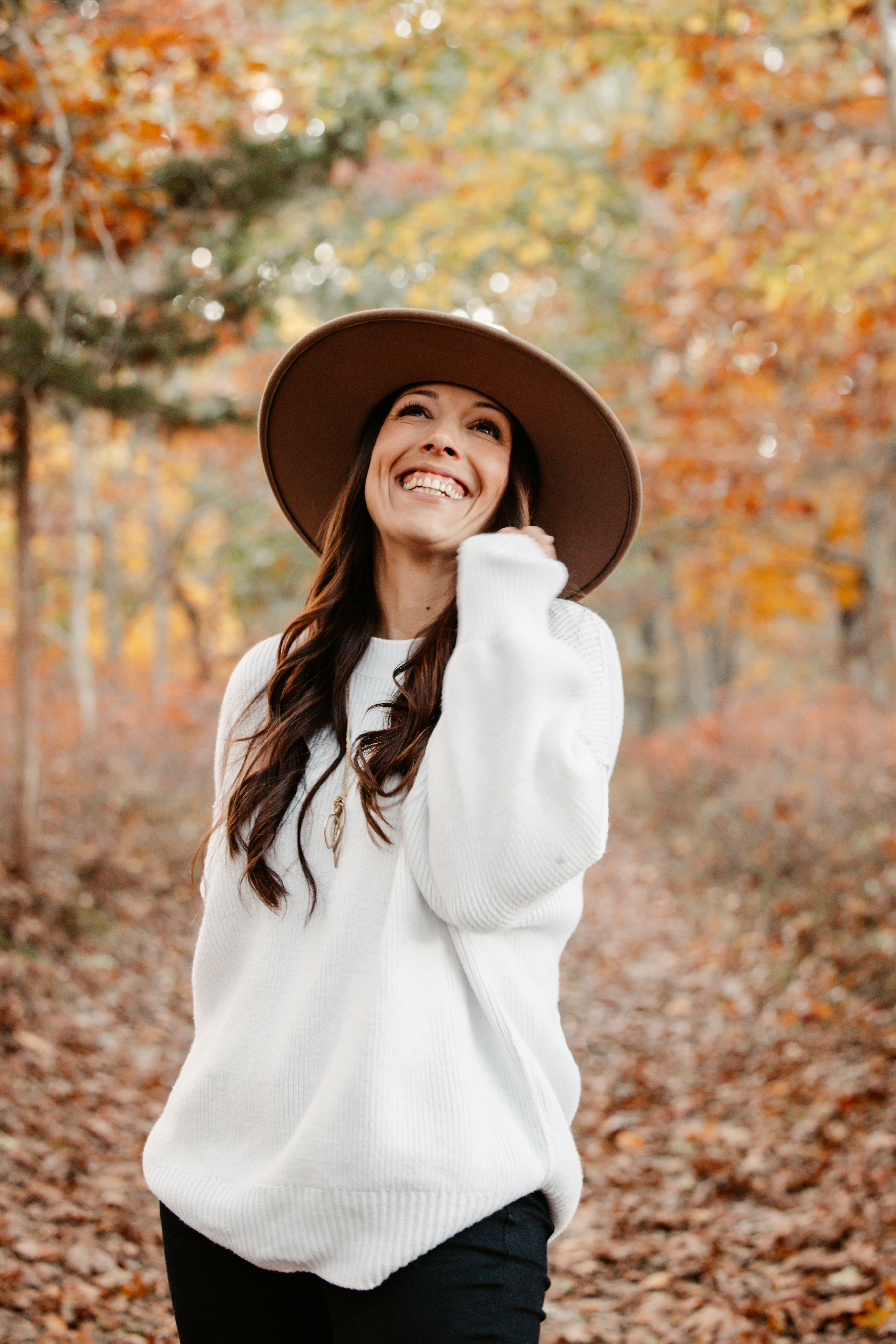 A smiling woman in a white sweater and brown hat in an autumn forest