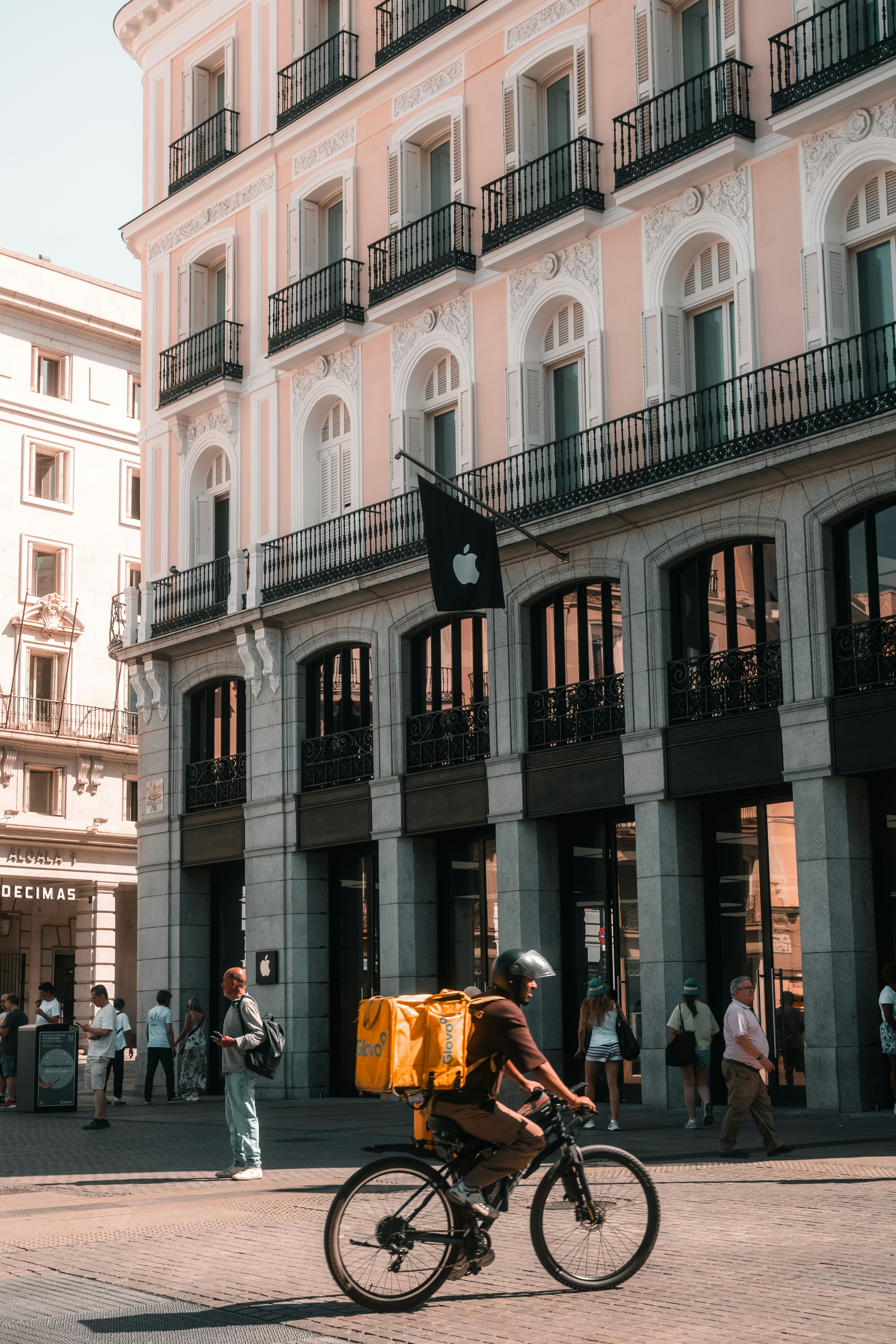 A delivery cyclist with a yellow backpack riding past a pink building in Madrid