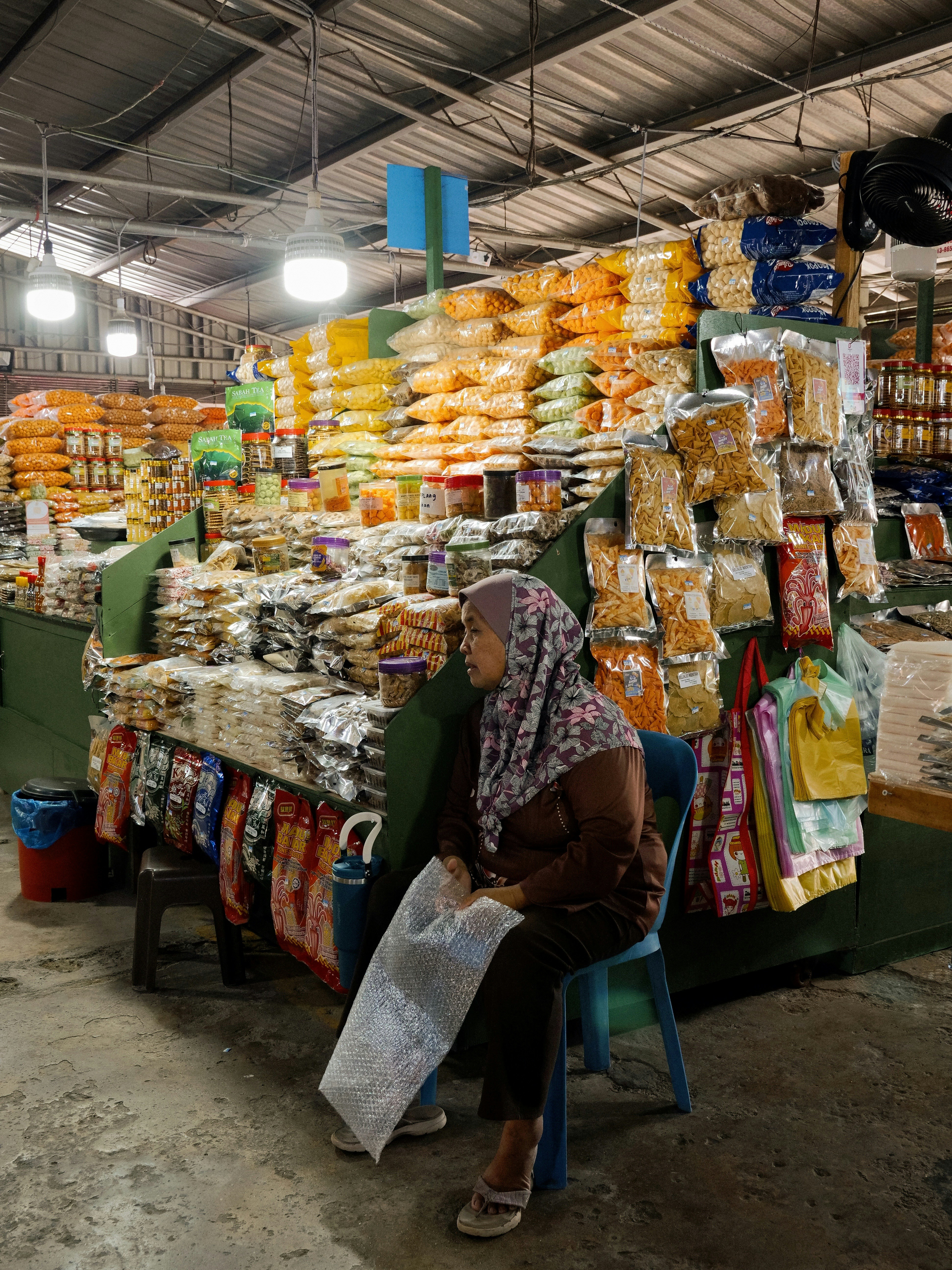 Une femme en hijab fleuri assise sur une chaise bleue à un stand de snacks