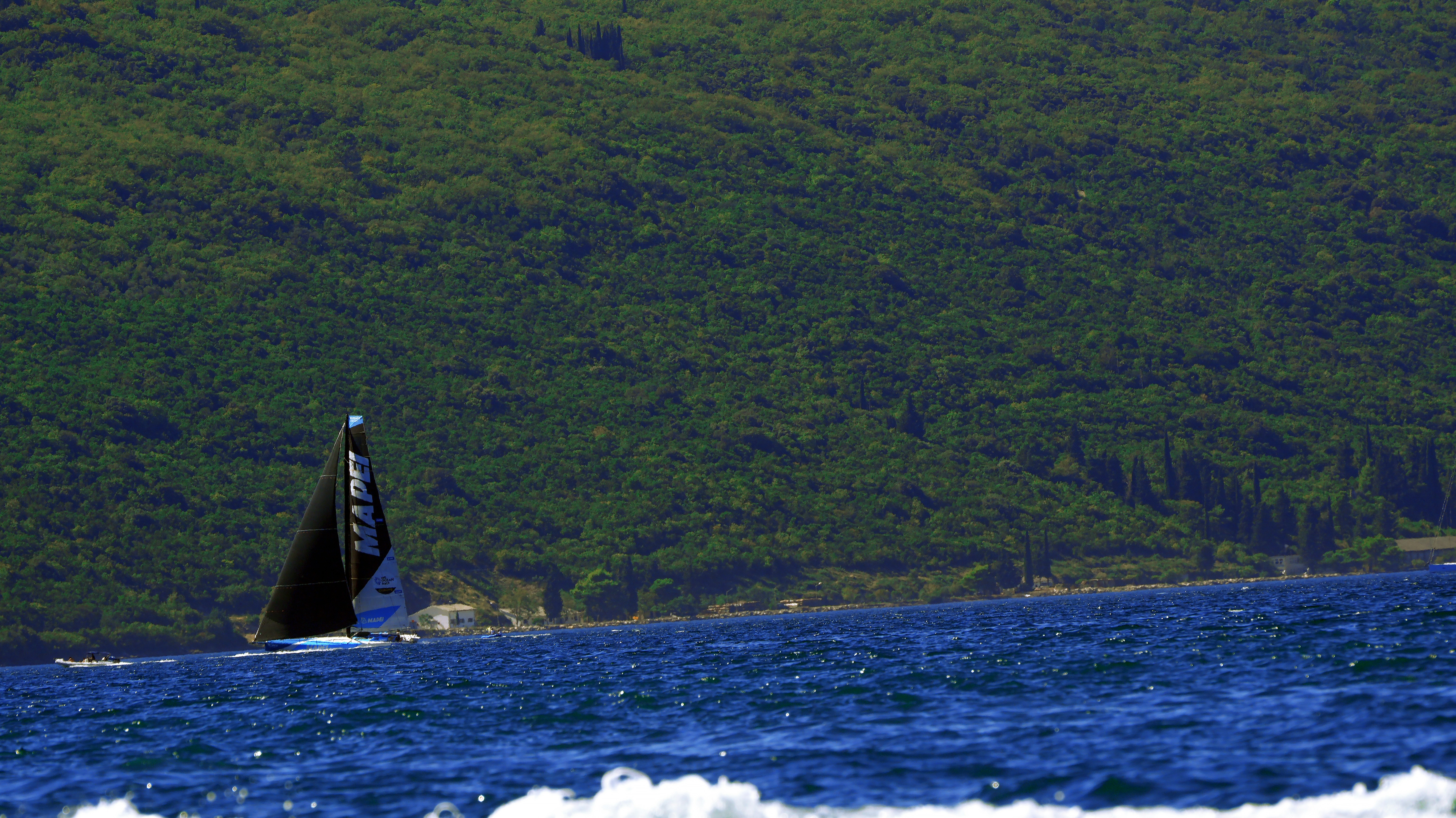 A sailboat with a black sail on blue water before a forested mountain