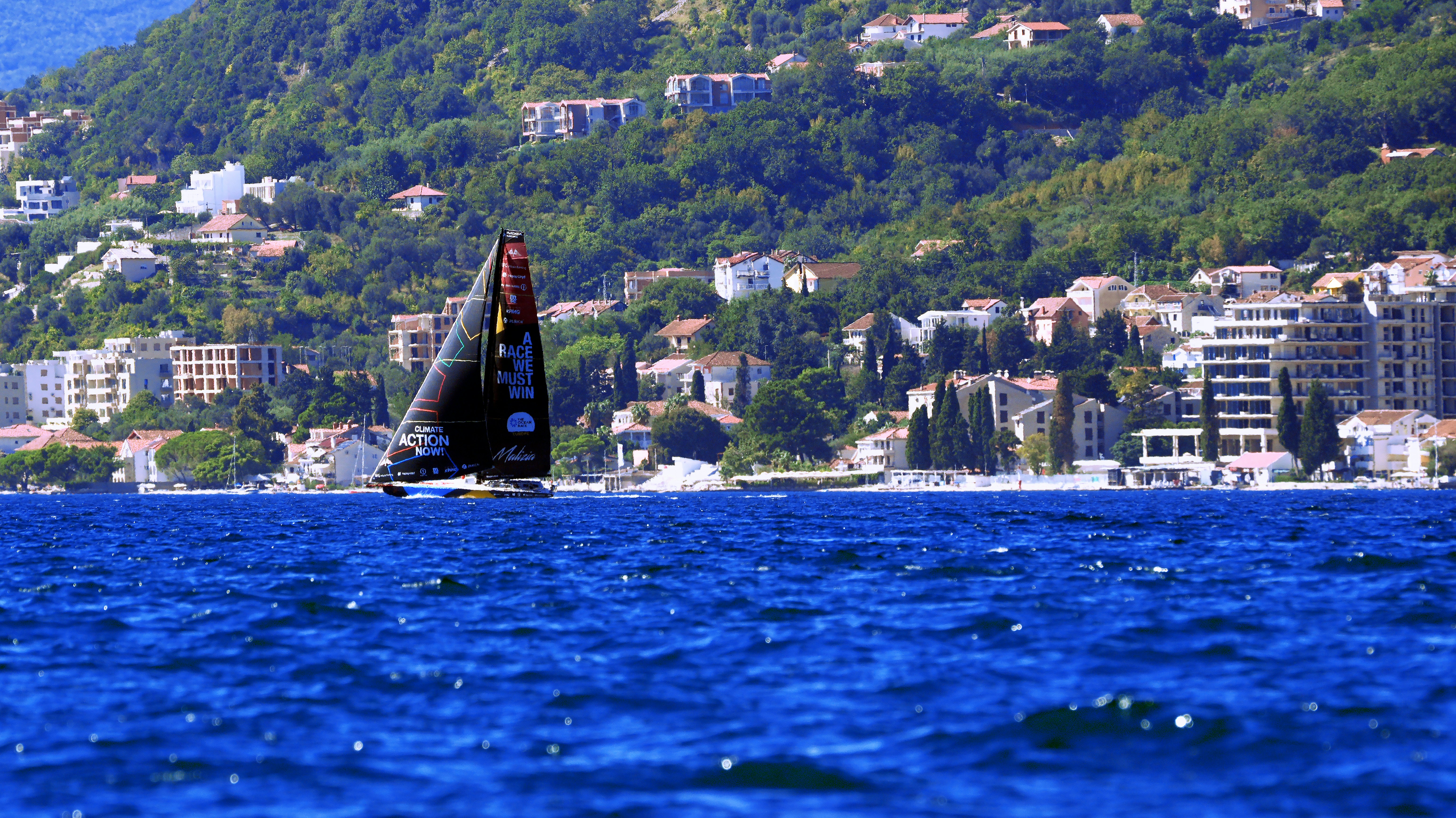 A sailboat with a black sail on blue water before a hillside town