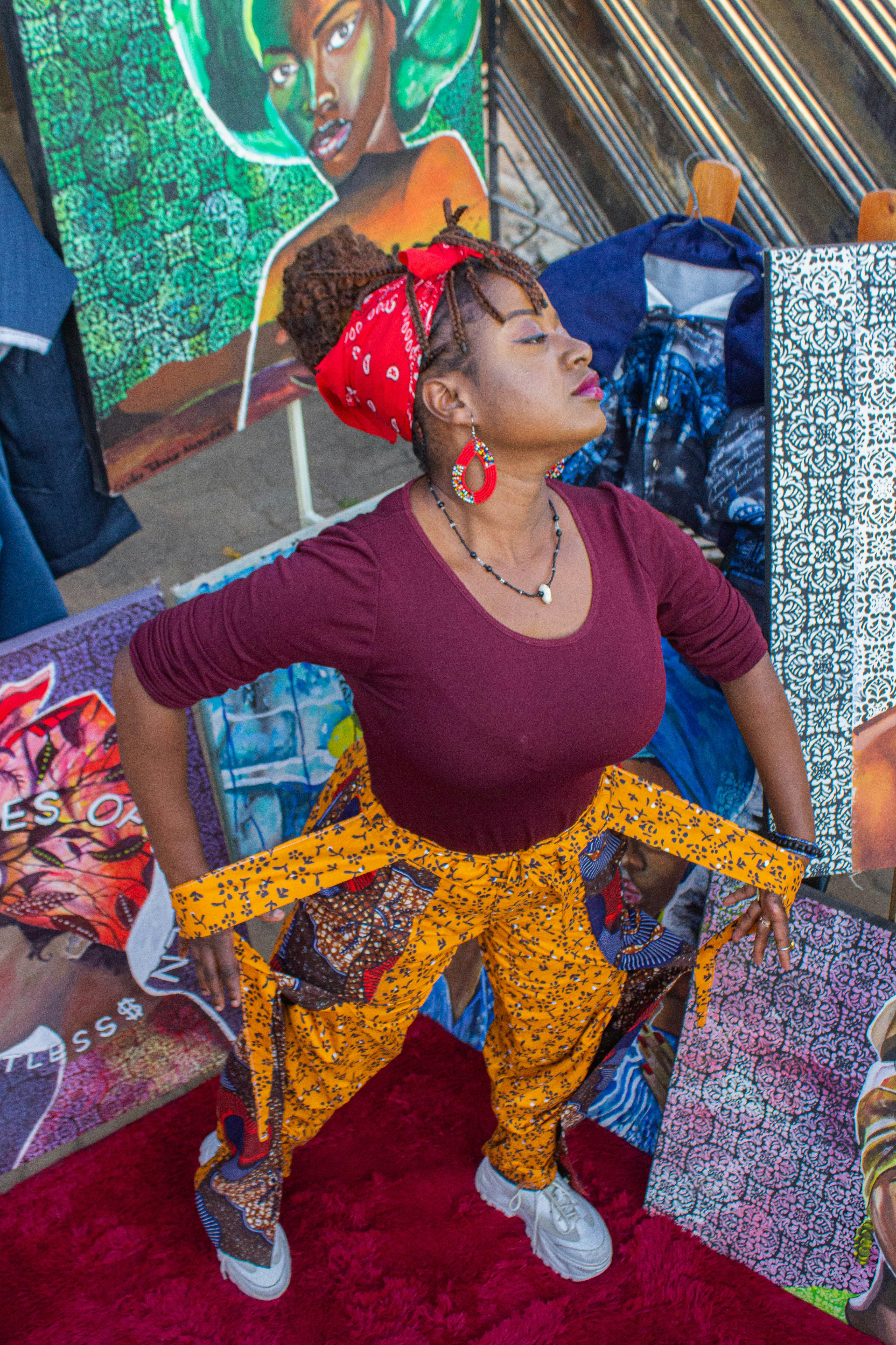 A woman in a maroon top and patterned yellow trousers standing among colorful paintings