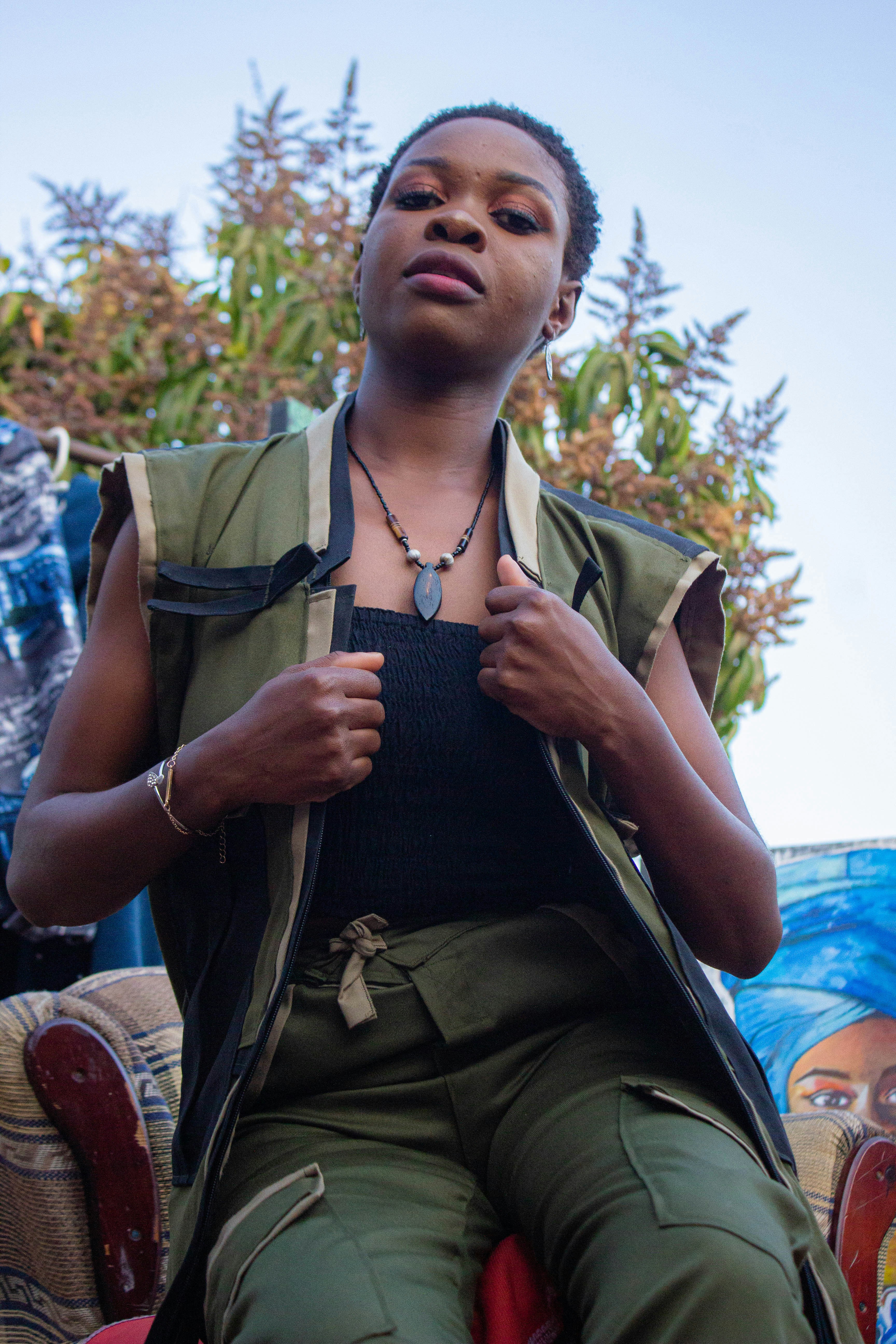A woman in an olive green vest and pants wearing a dark pendant necklace