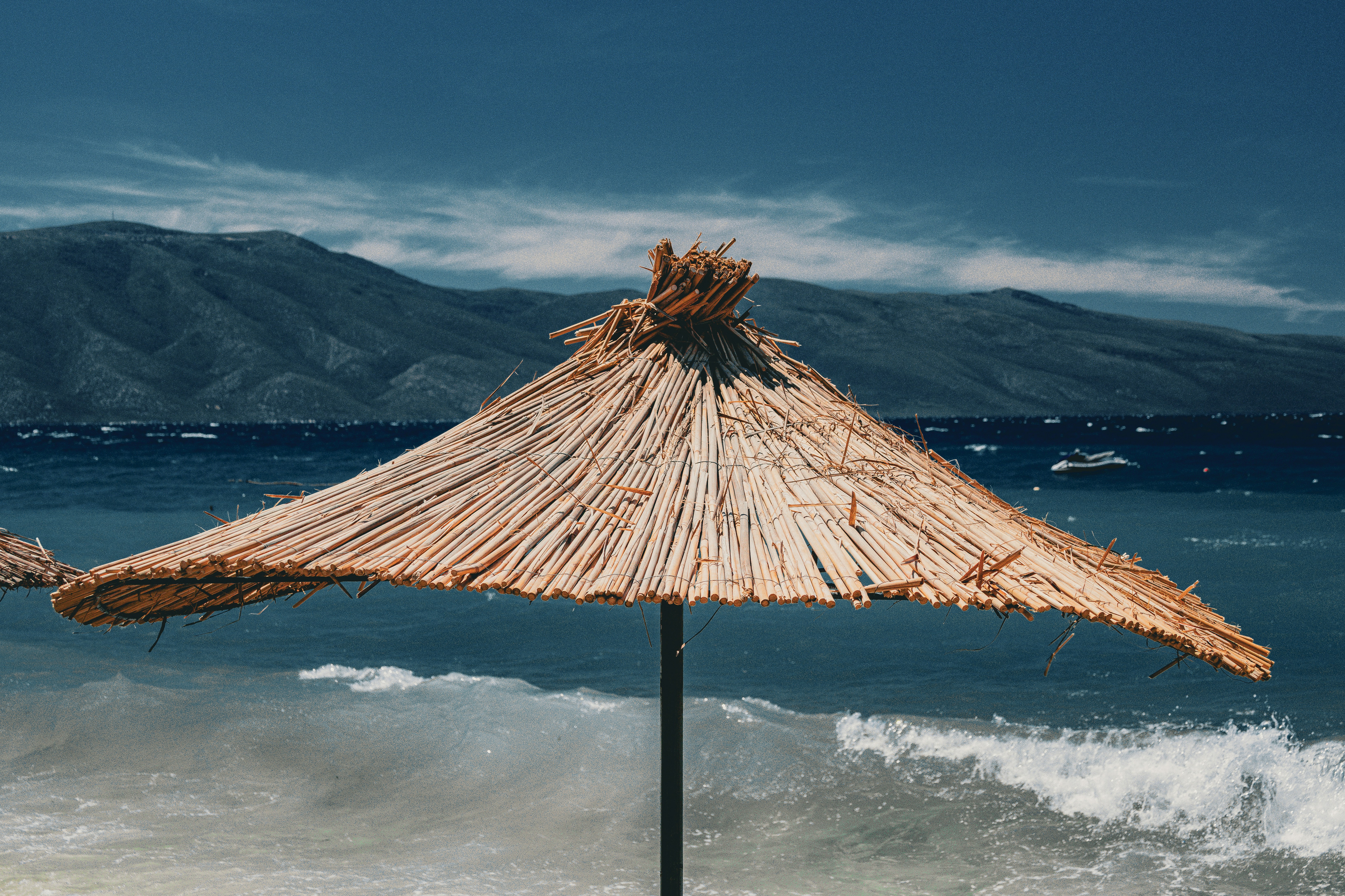 Thatched beach umbrella in strong winds over rough waves with distant mountains, suggestive of October weather in Albania