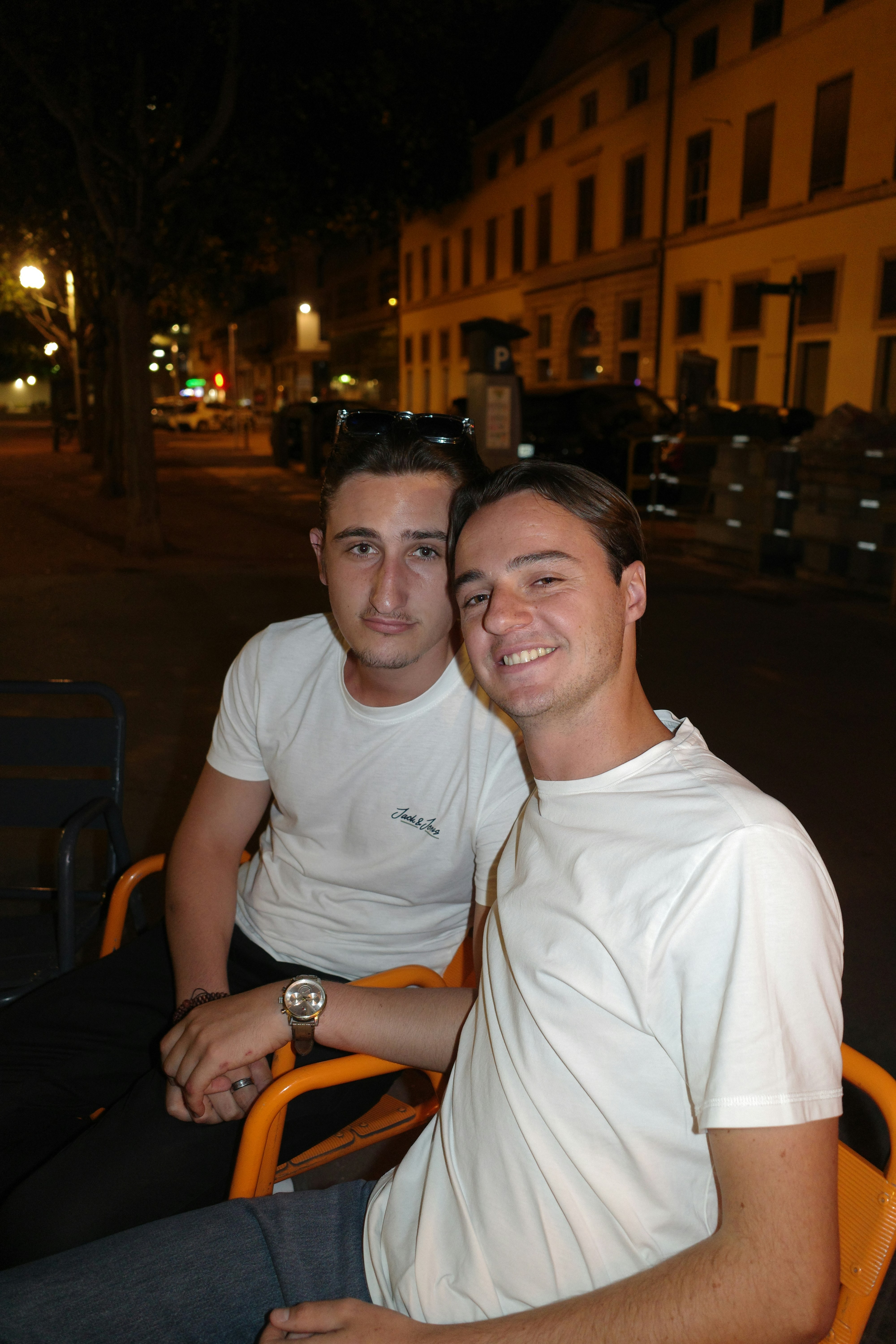 Two men in white t-shirts sitting on orange chairs on a city street