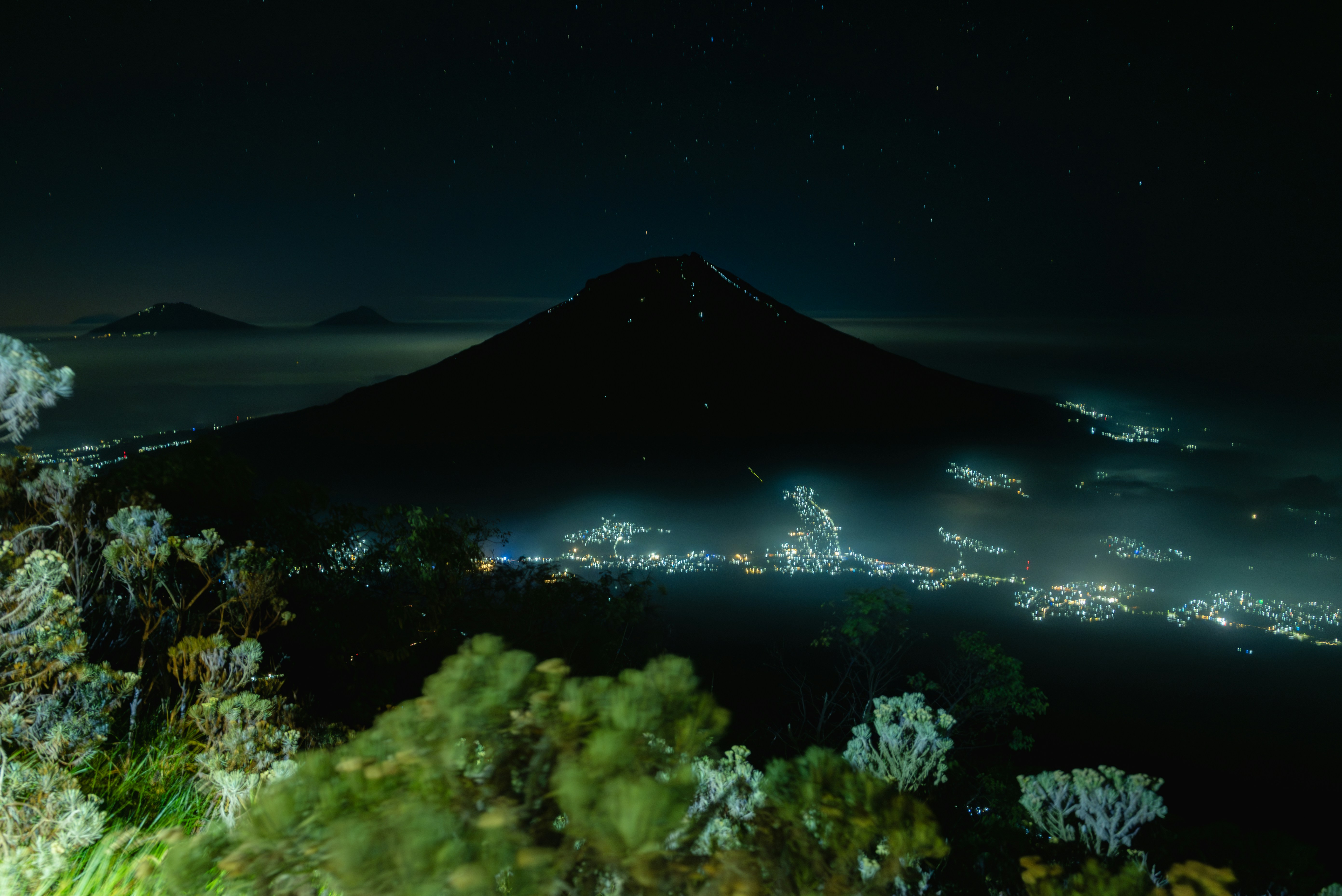 A dark mountain silhouette above a glowing town at night under a starry sky