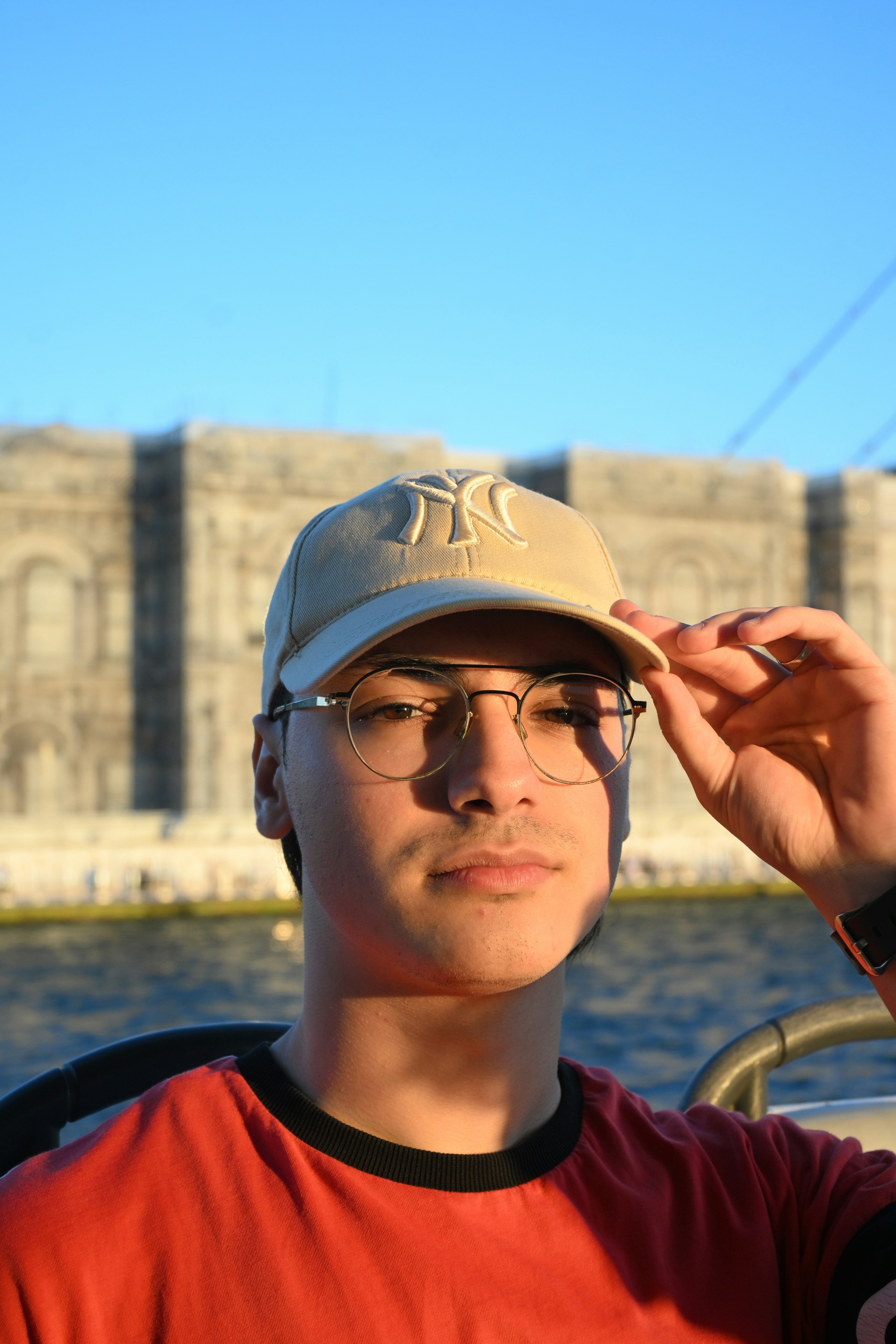 Una persona con una gorra de béisbol beige y gafas contra un edificio borroso frente al agua