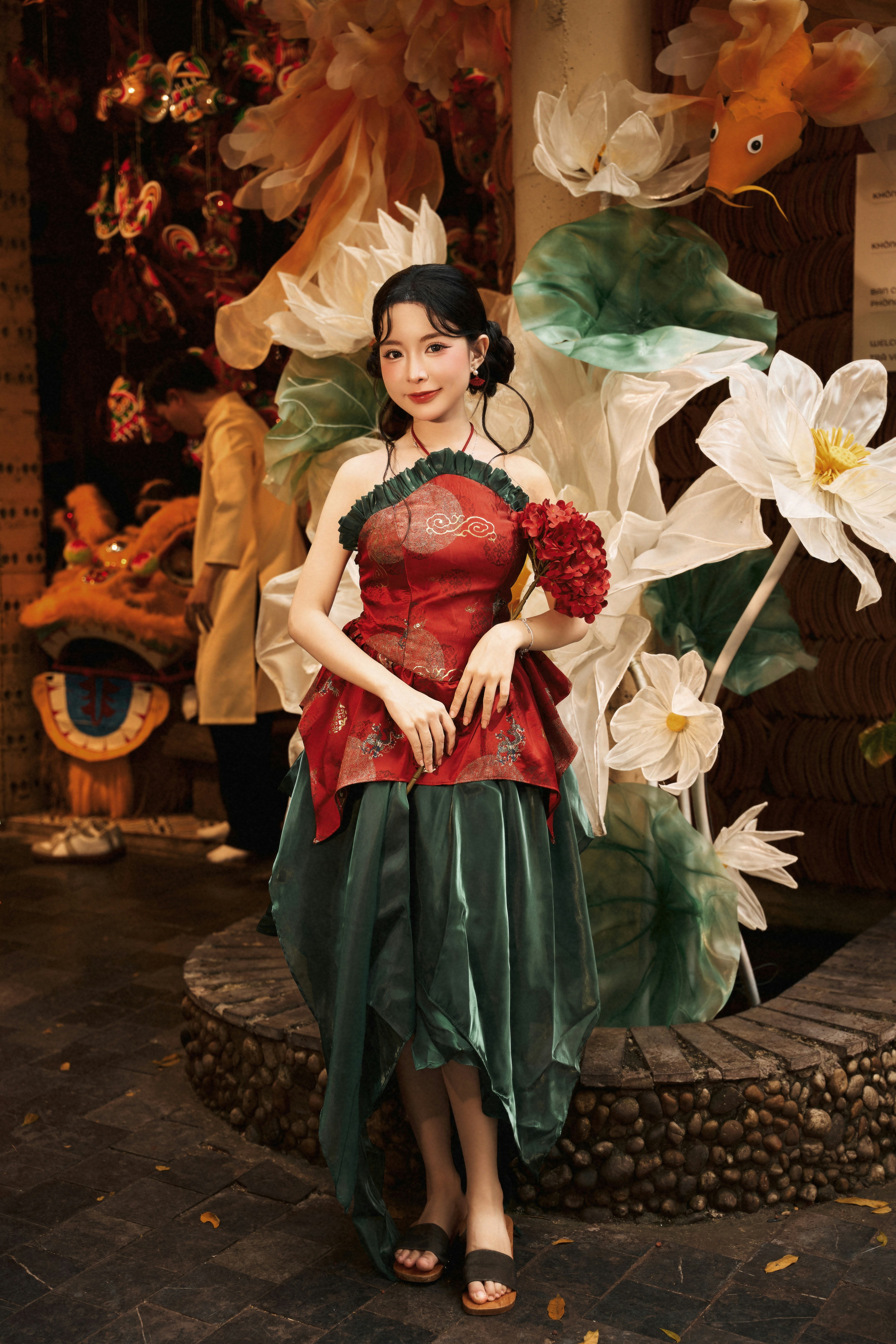 A woman in a red and green traditional dress holding red flowers near lotus decorations