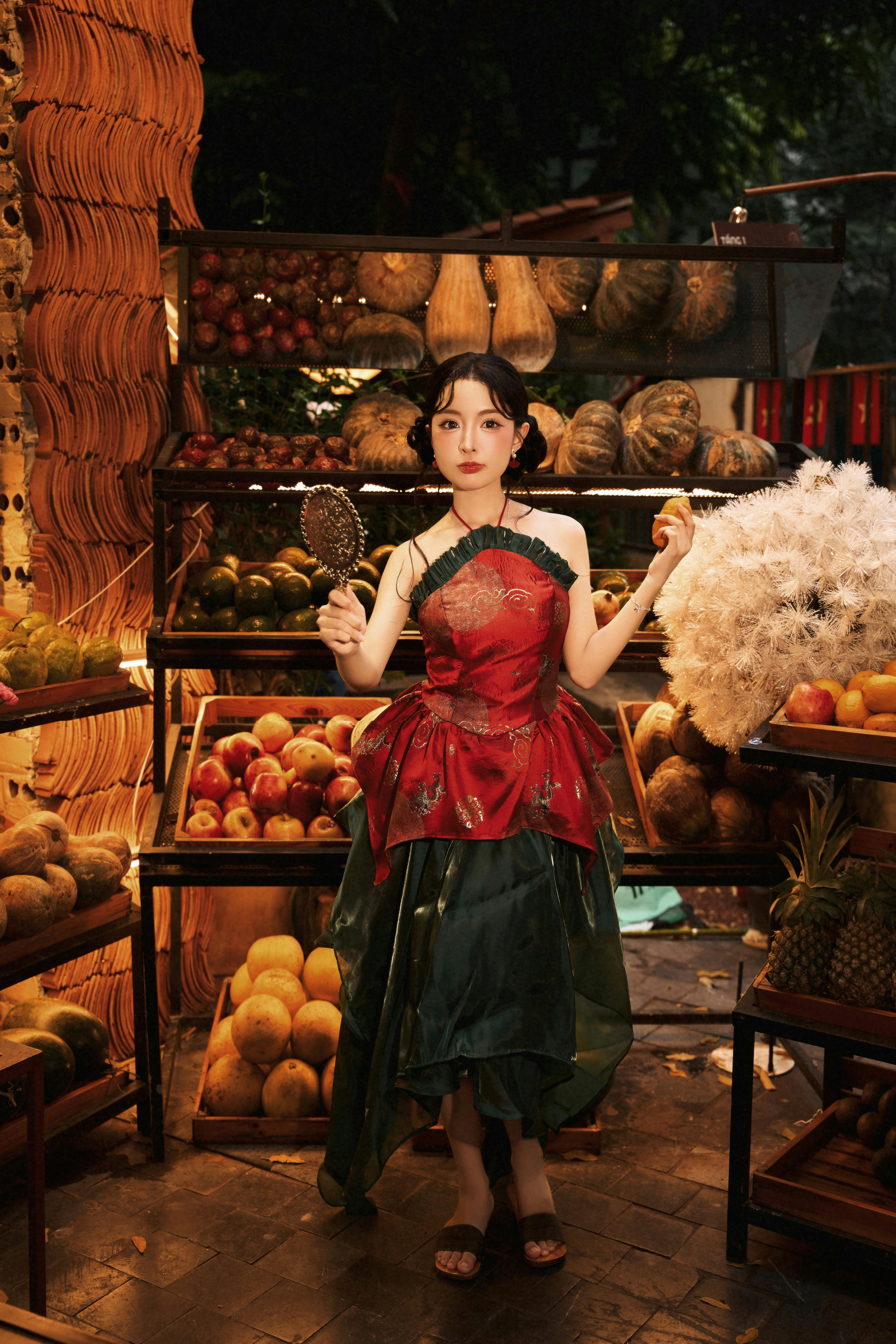A woman in a red and green dress holding a mirror beside fruit shelves