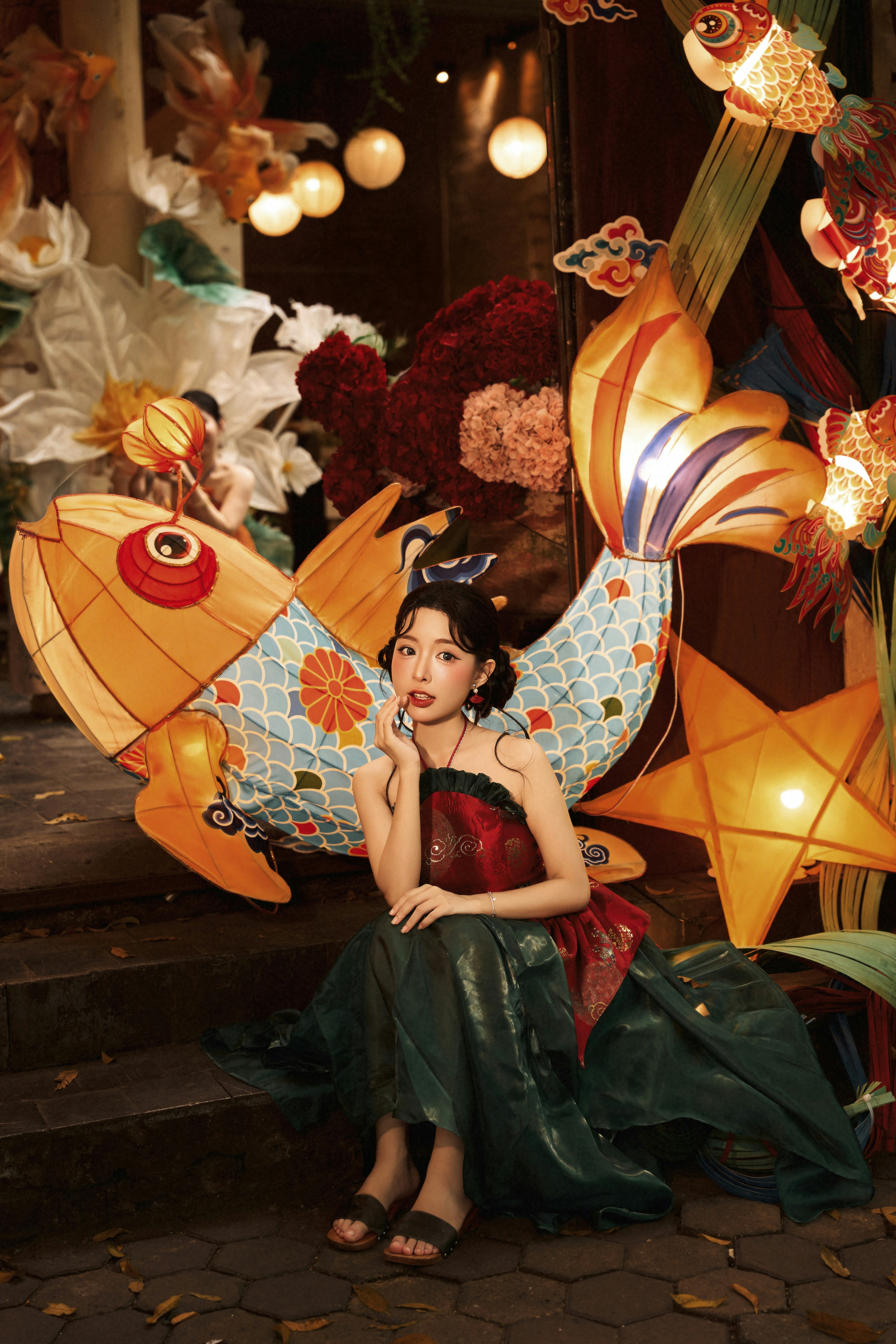 A woman in a traditional red and green dress sitting among illuminated fish lanterns