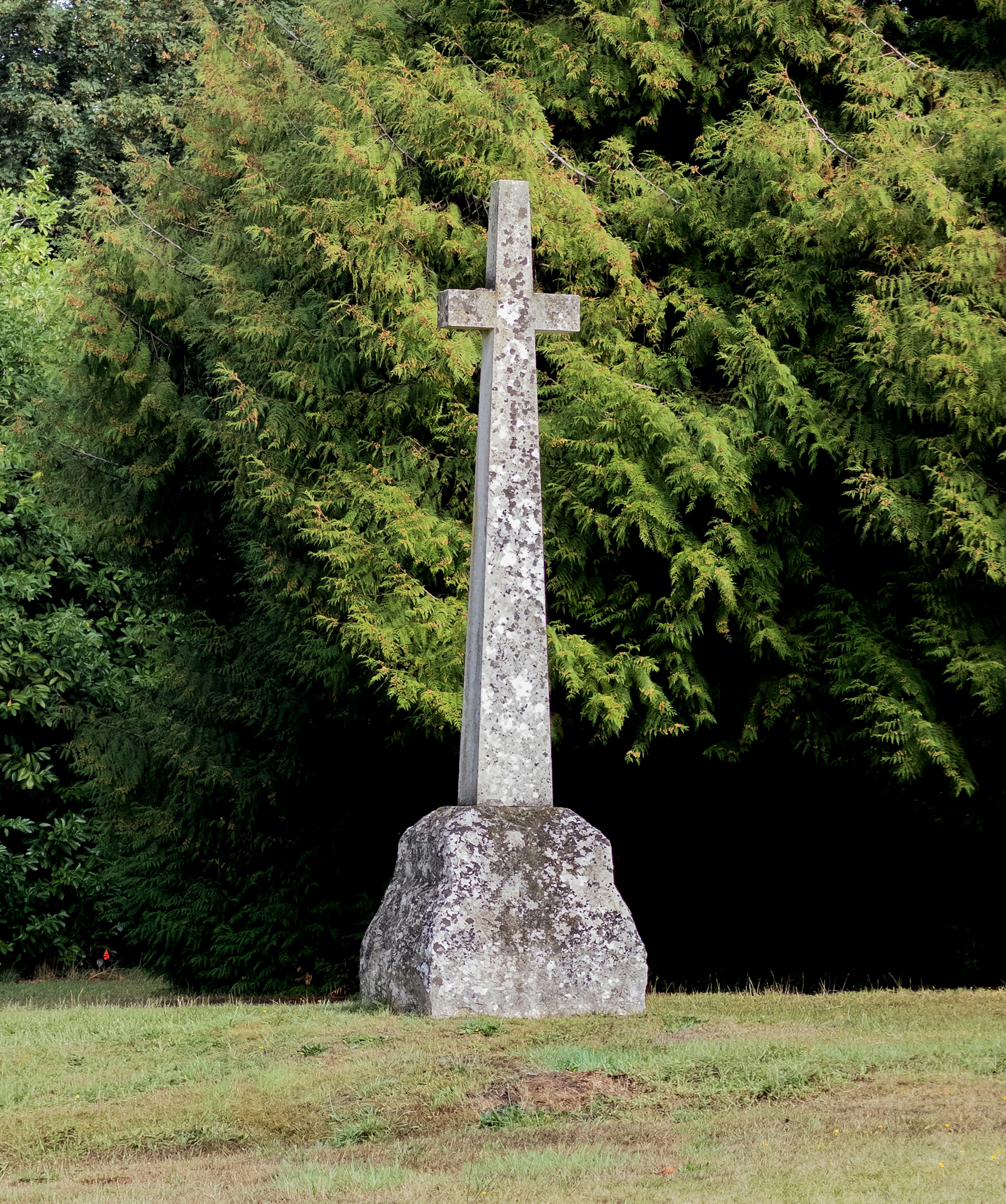 A weathered stone cross on a rough base before dense green evergreen trees