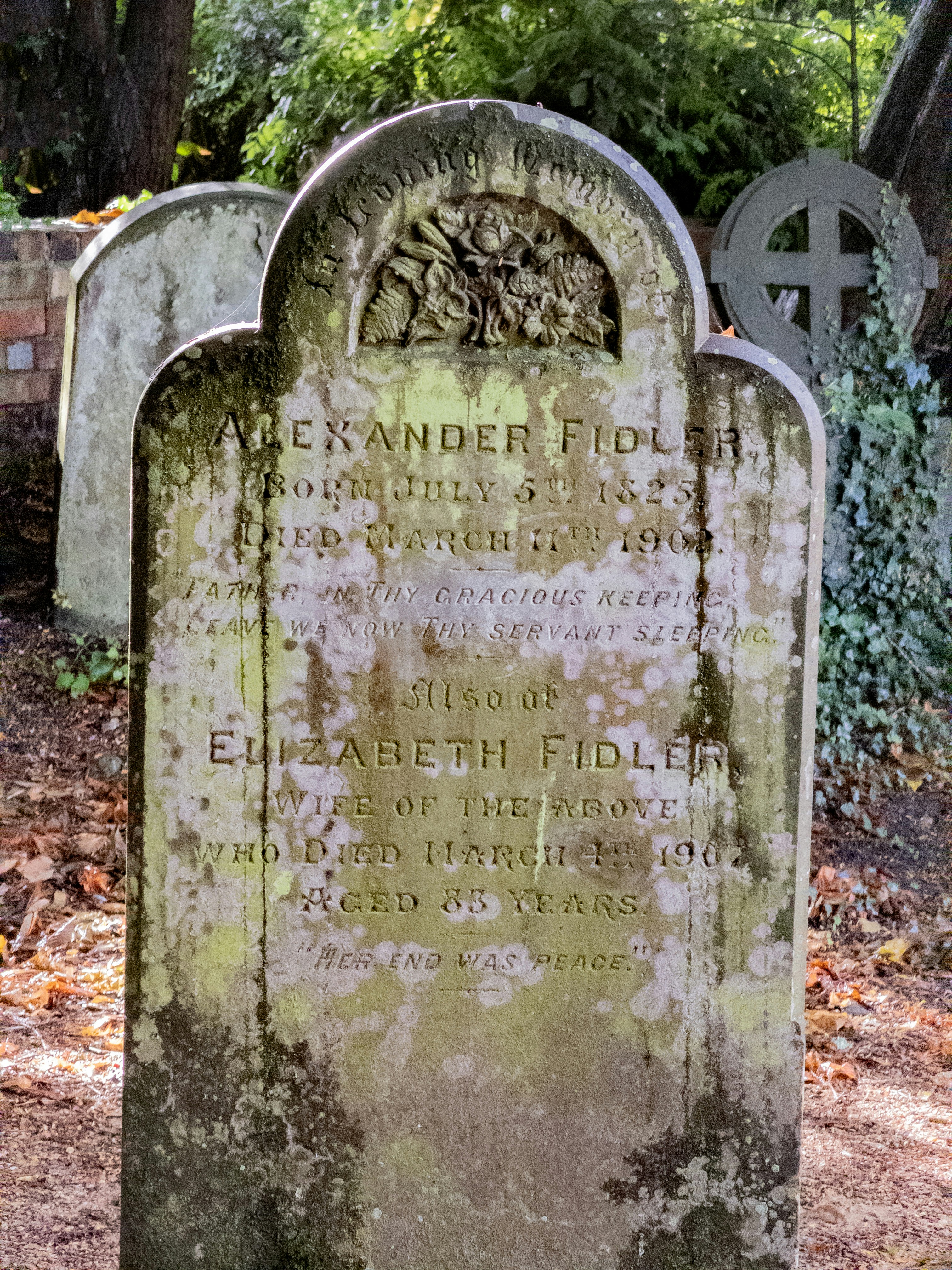 A weathered stone headstone with moss in a cemetery with other grave markers