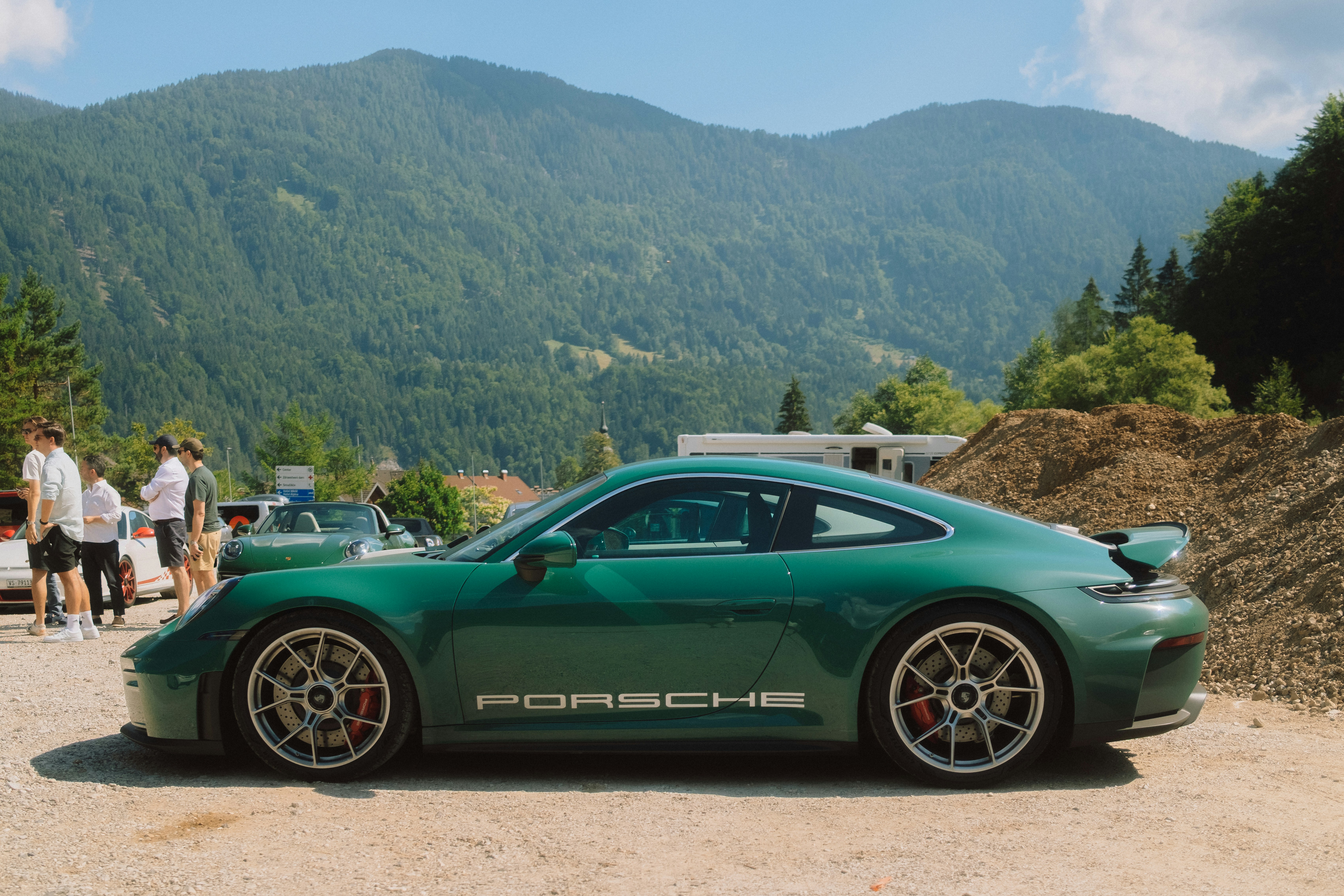 A green Porsche sports car parked on a gravel lot before forested mountains