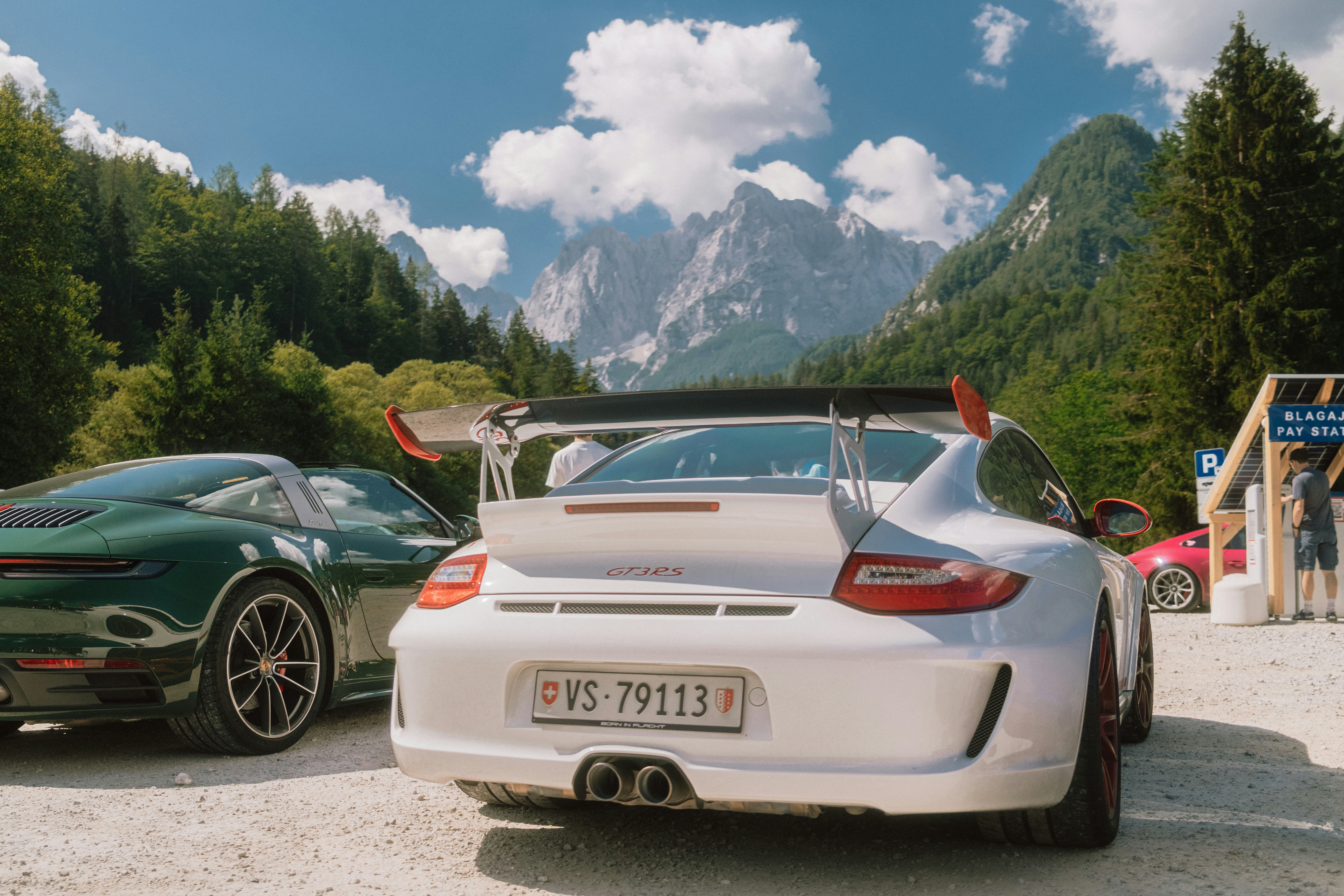 A white Porsche GT3 RS and a green sports car parked before mountain peaks