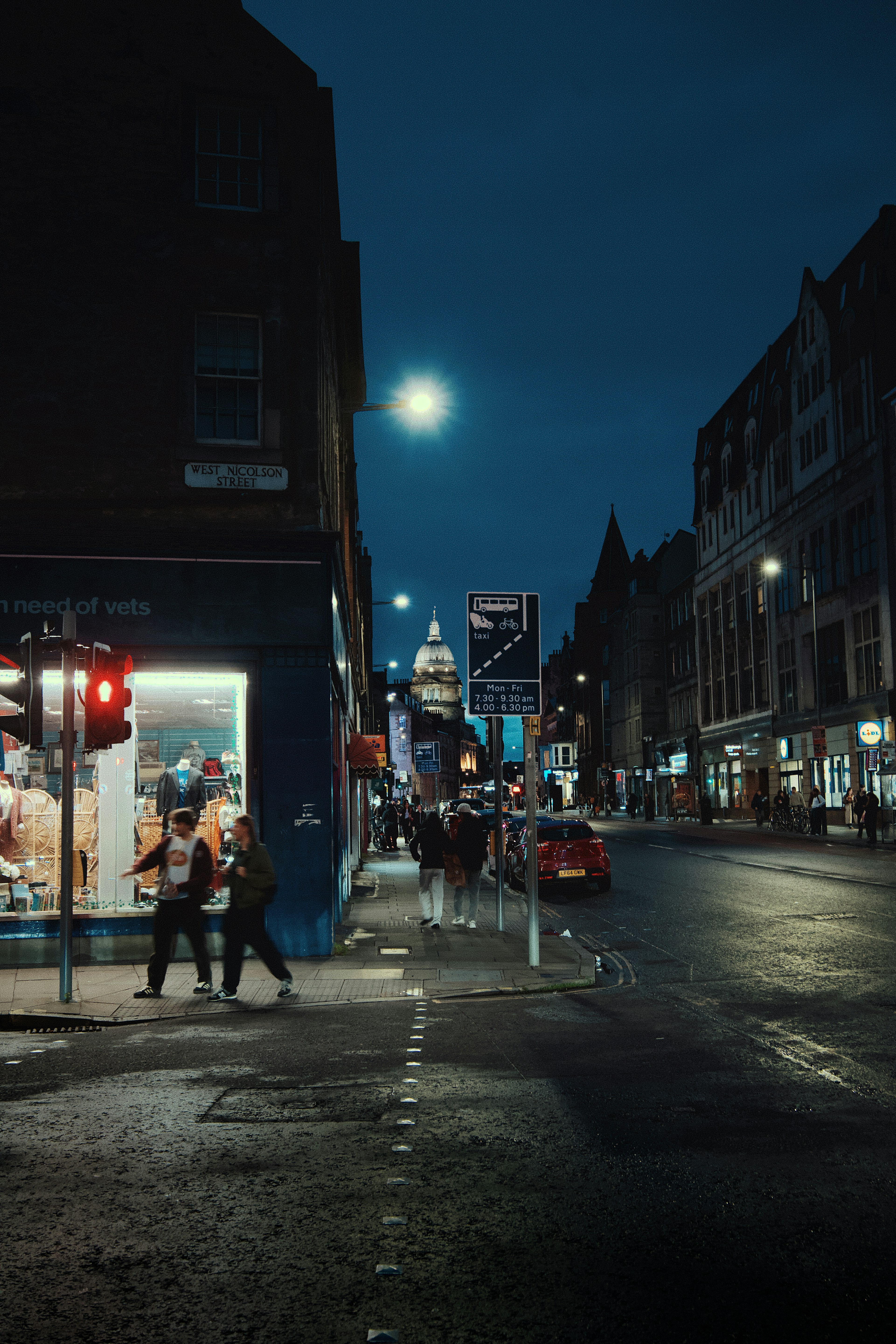 People walking along West Nicolson Street in Edinburgh at night, under streetlights, with shops lit up