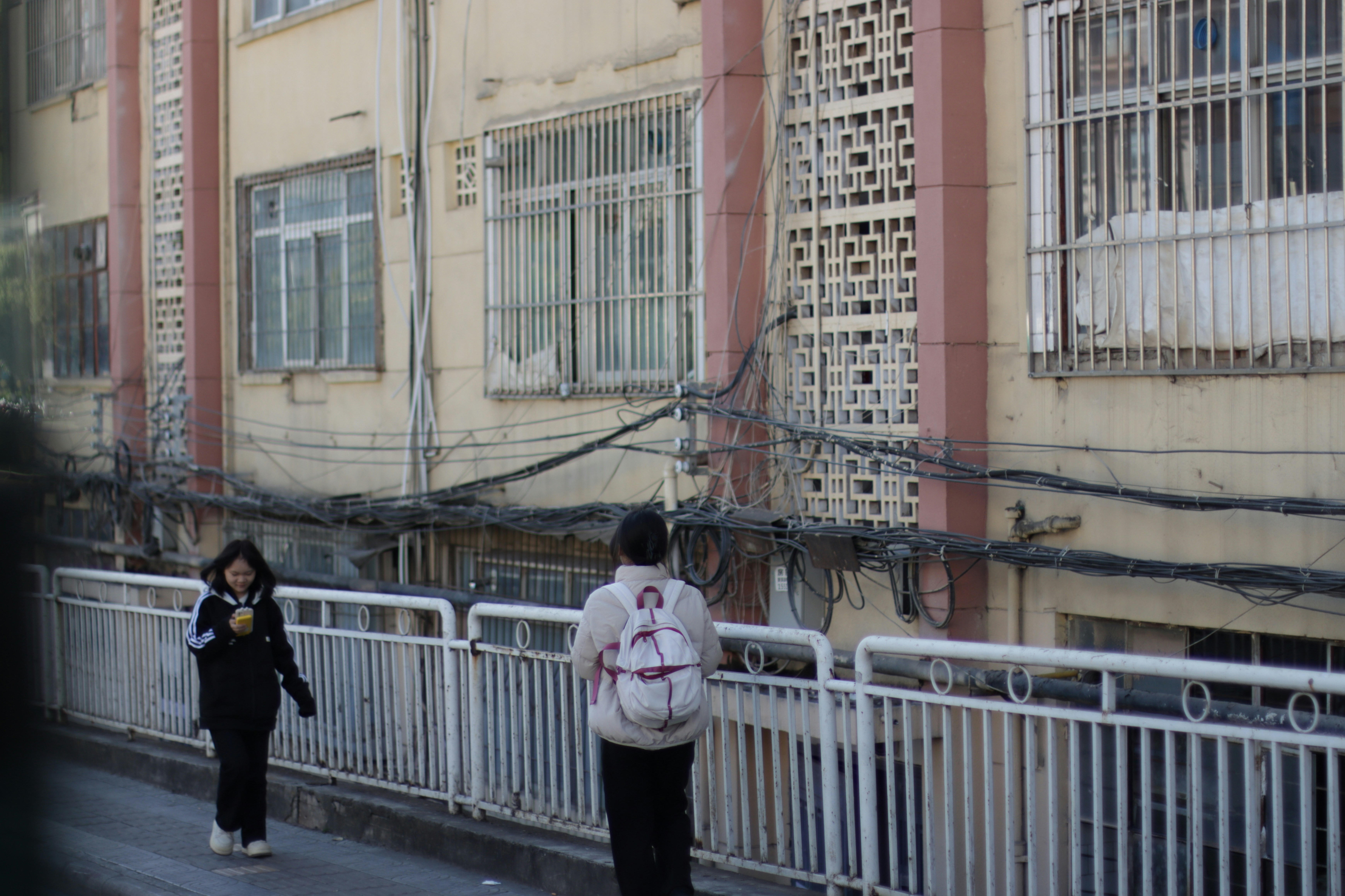 Two people walking along a sidewalk beside a building with tangled overhead wires