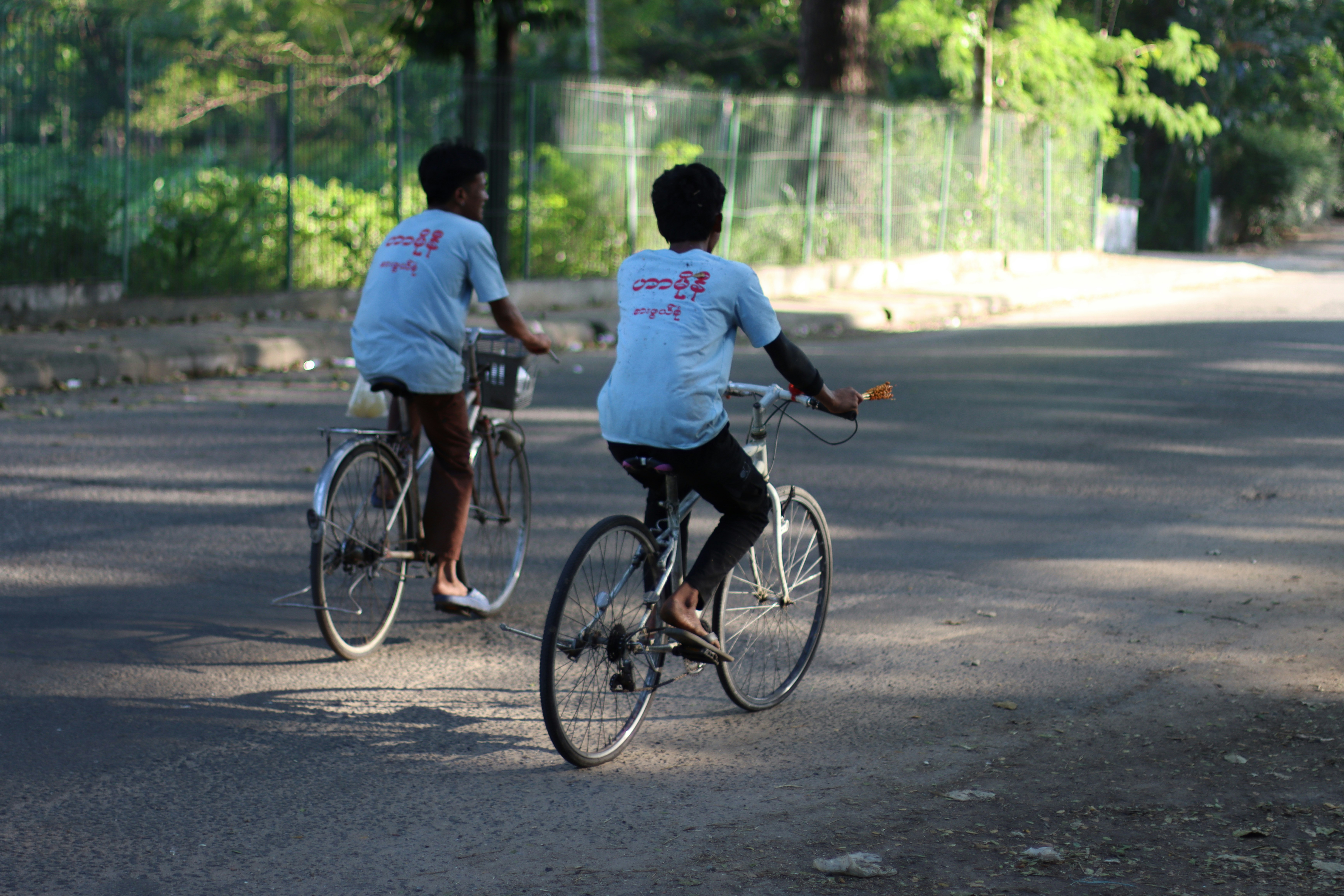 Two people in light blue shirts cycling on a paved road in sunlight