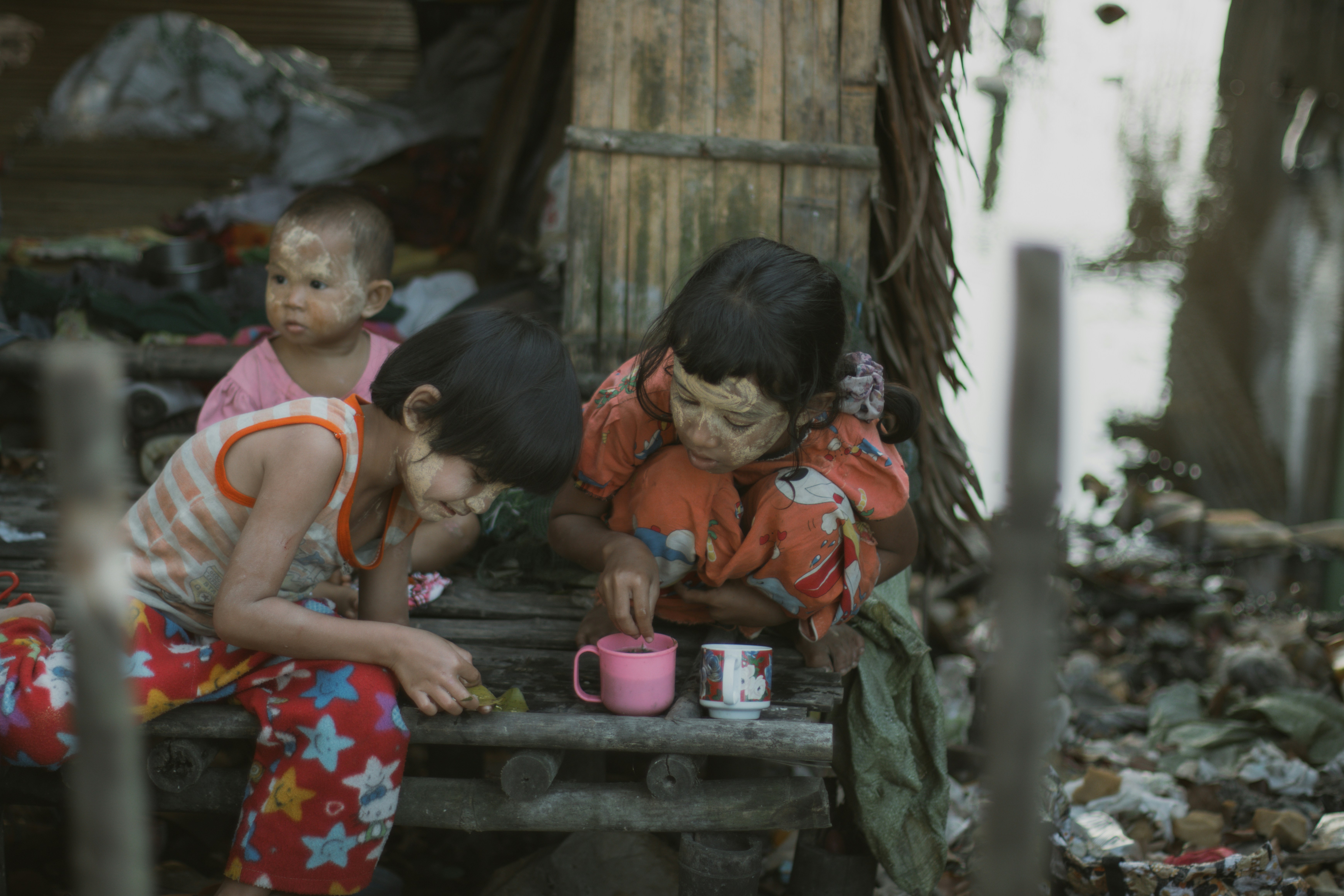 Three children with thanaka paste on their faces sitting on a bamboo platform