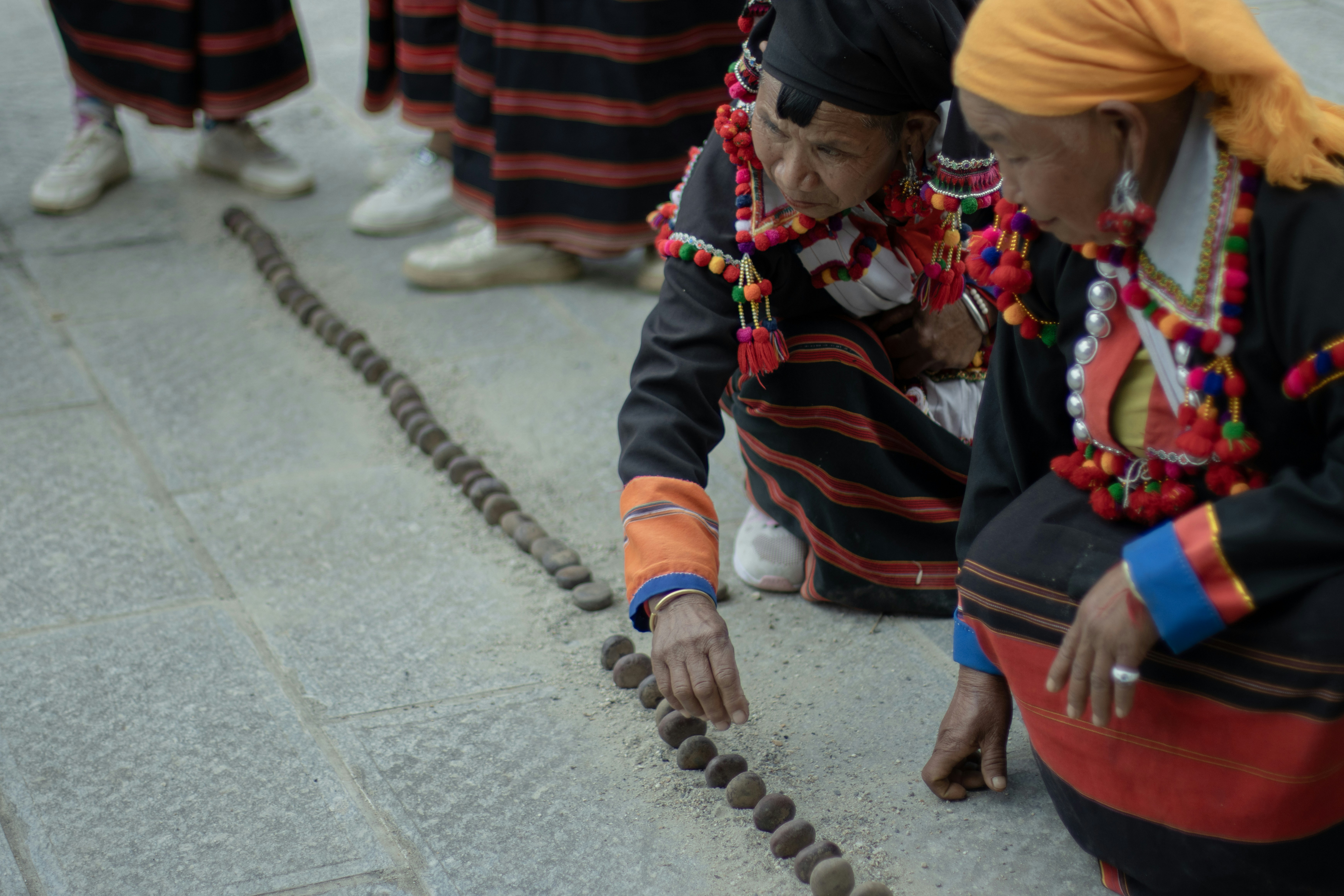 Two women in traditional clothing placing small stones in a line on stone pavement