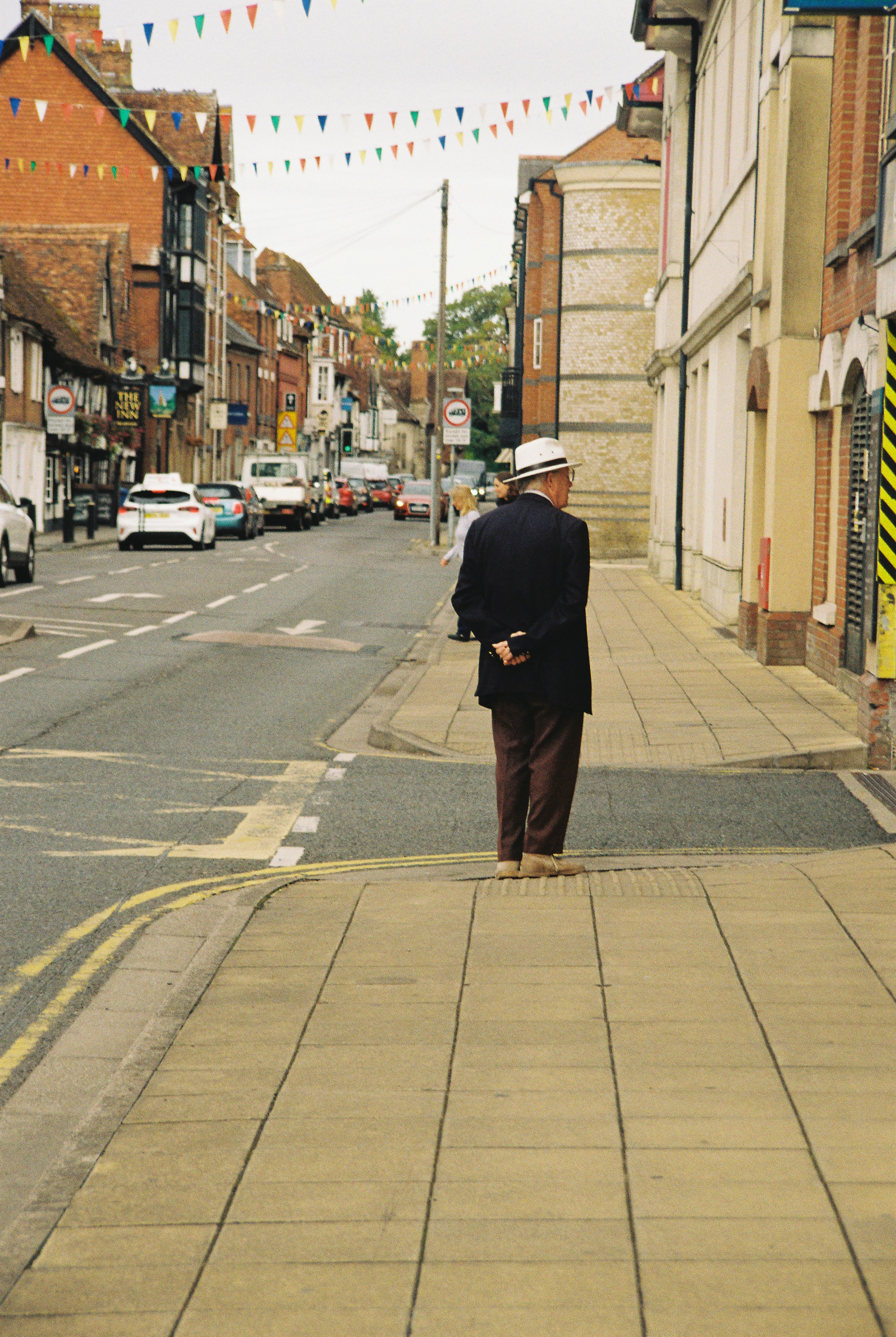 An older man in a hat and blazer on a sidewalk with bunting overhead