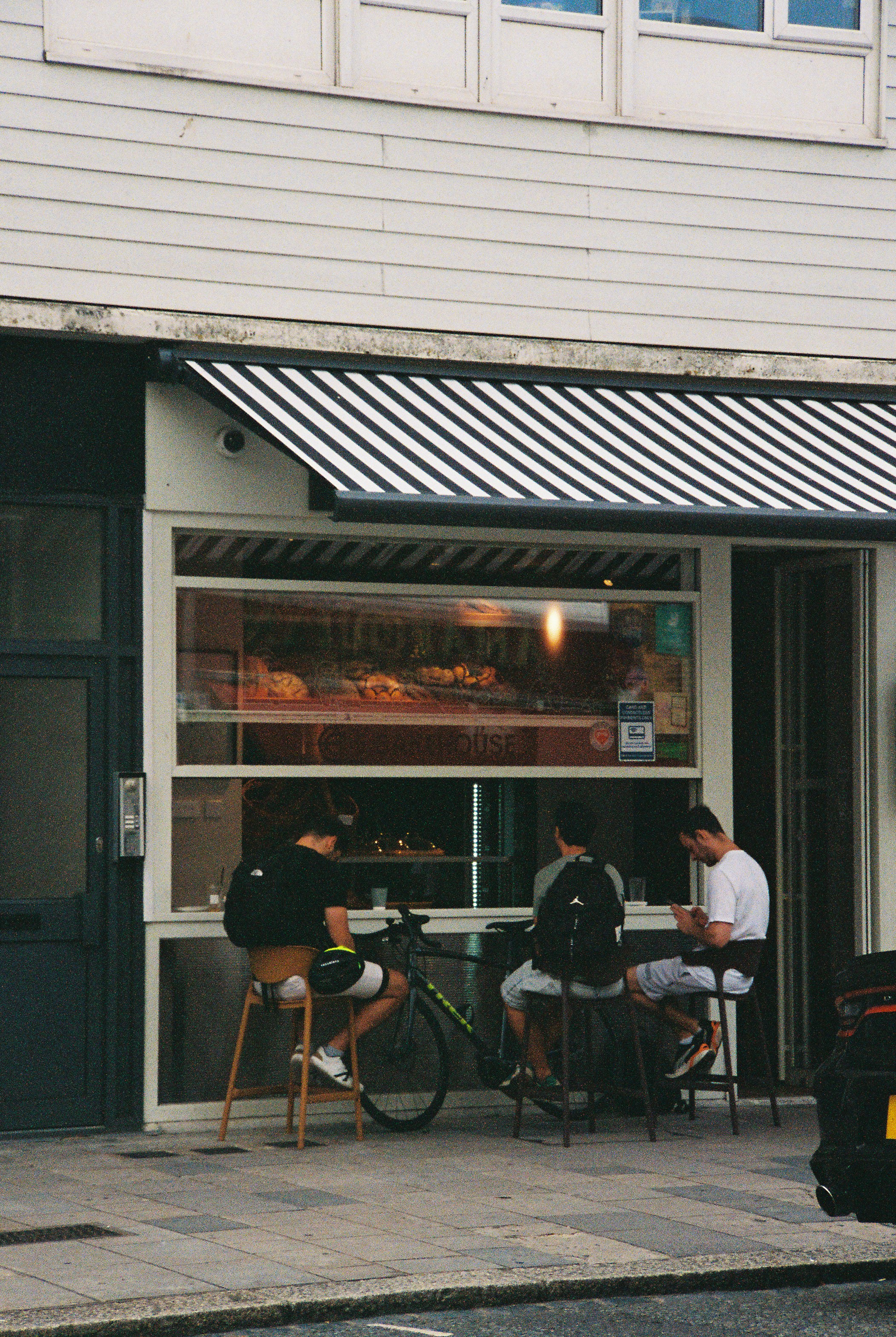 Three people sitting on stools at a cafe window with a striped awning
