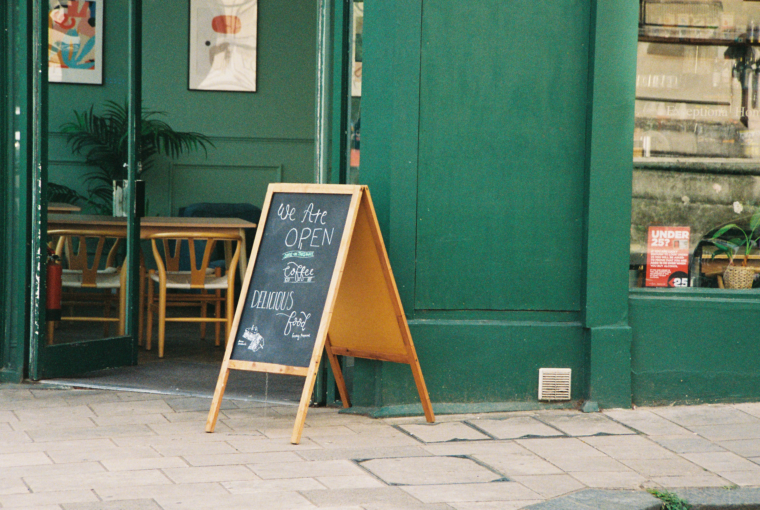 A chalkboard sign reading "We Are OPEN" on a sidewalk outside a green cafe