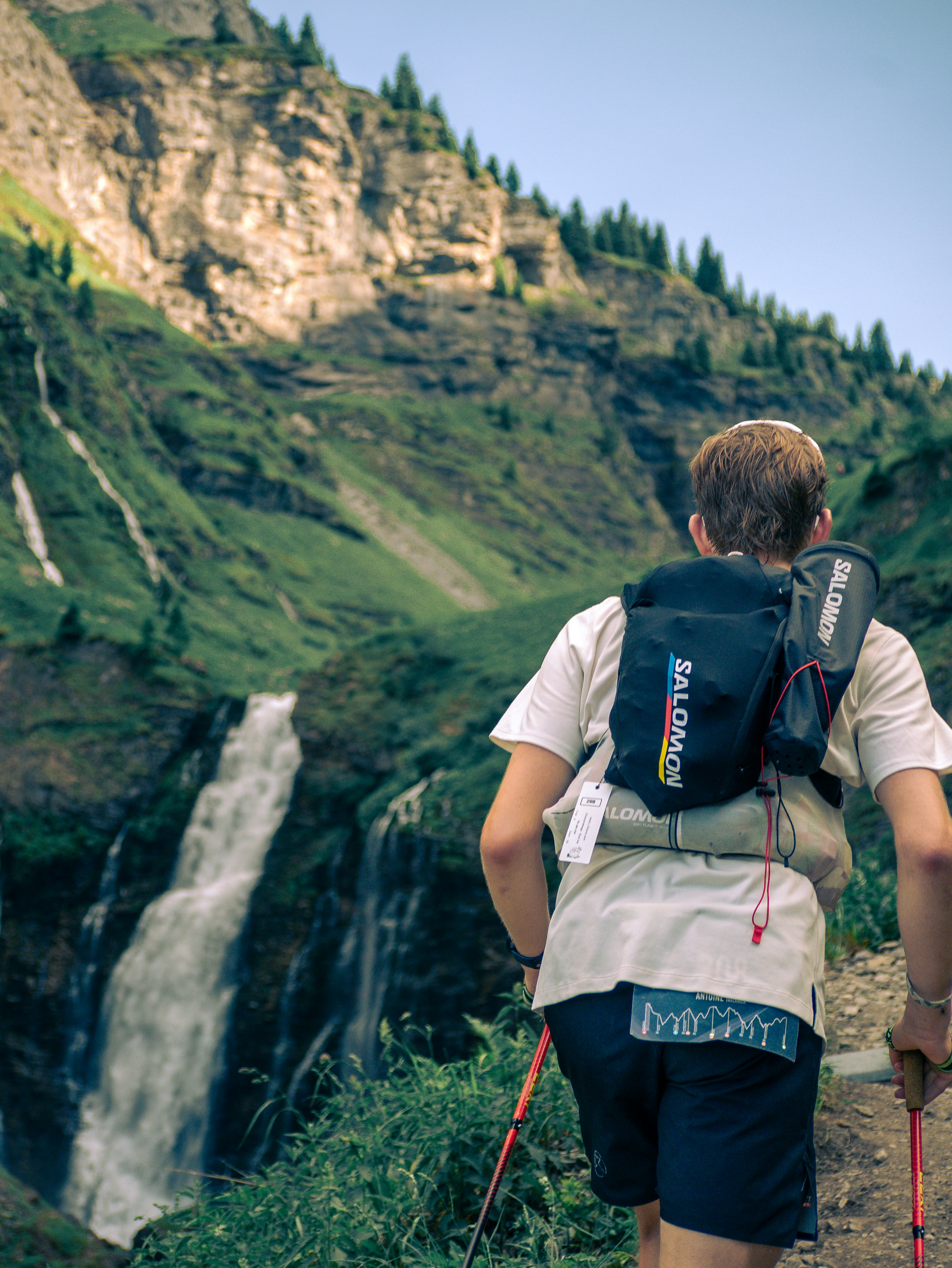 A person with a backpack and trekking poles hiking past a mountain waterfall