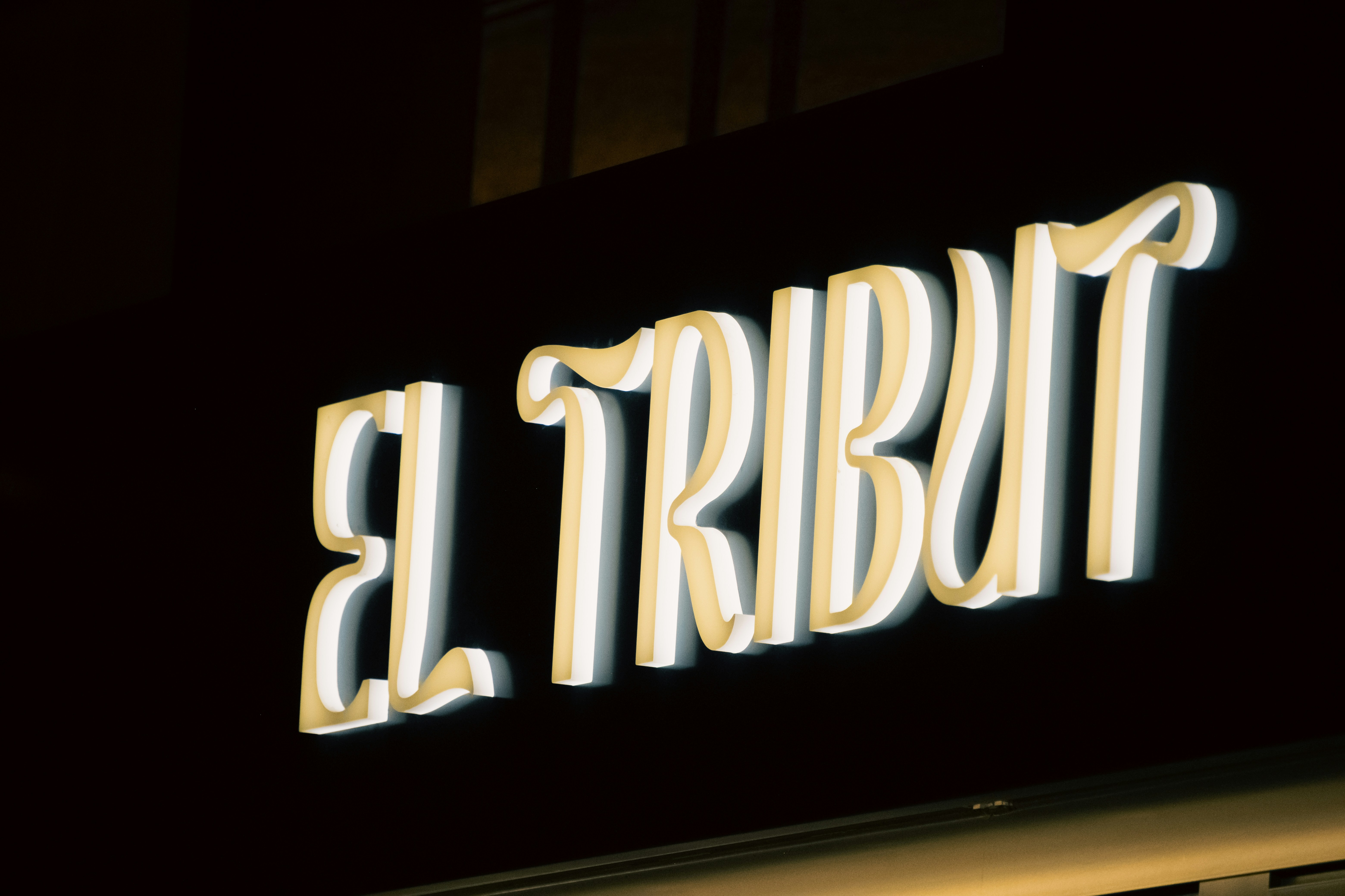 A glowing neon sign reading EL TRIBUT against a dark background