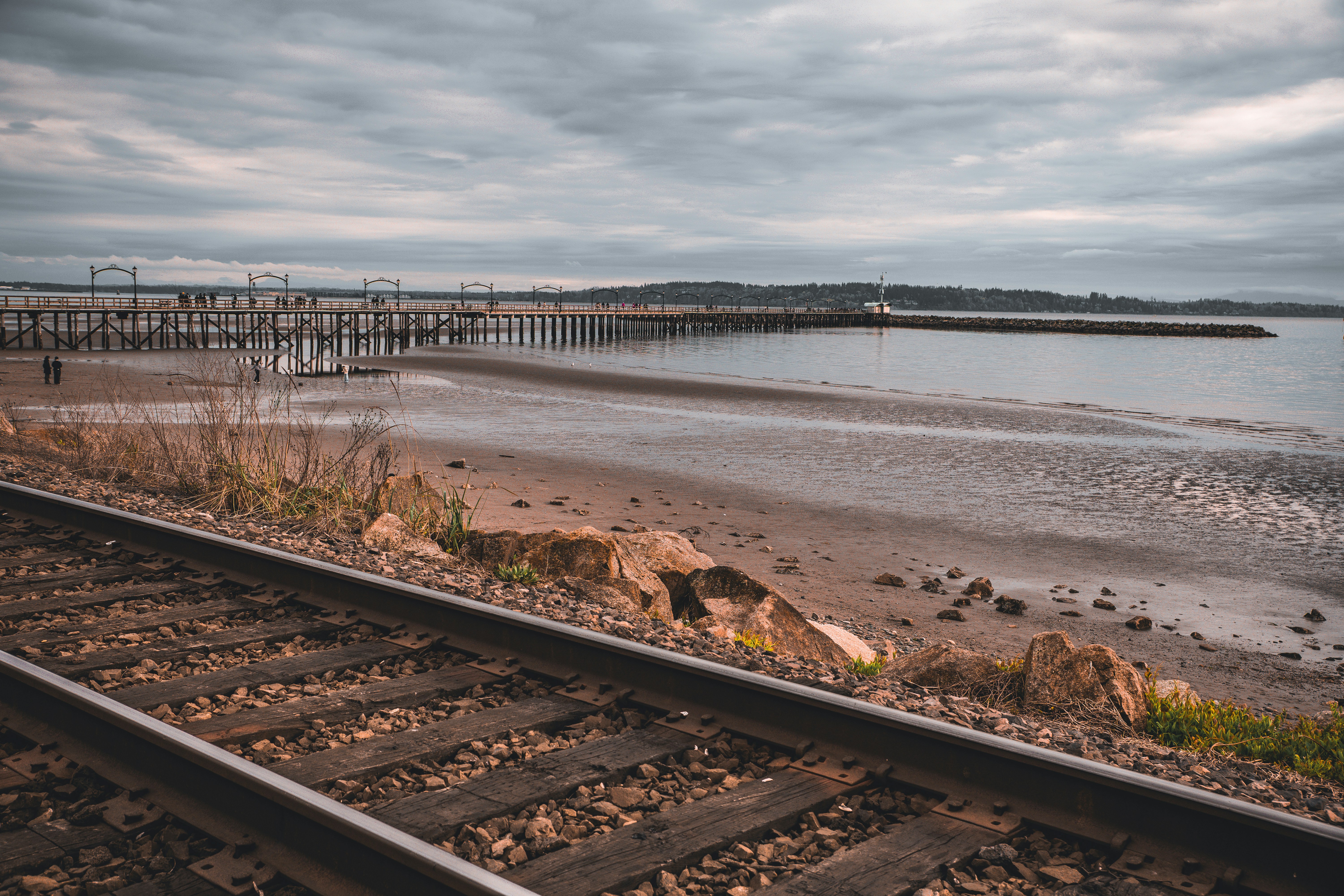 Railroad tracks running along a sandy beach with a long pier under cloudy skies