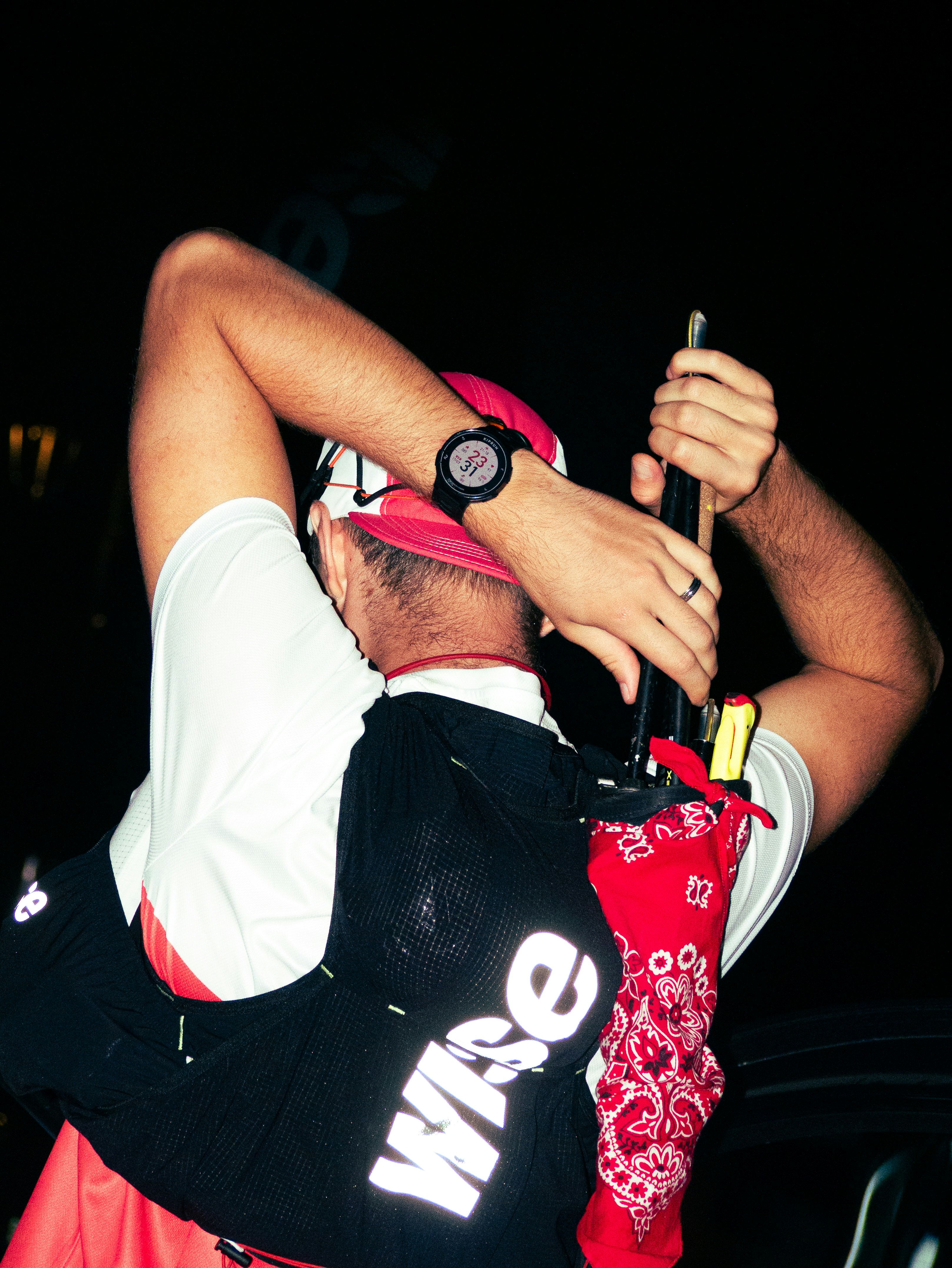 A person wearing a black running vest and red cap adjusting gear at night