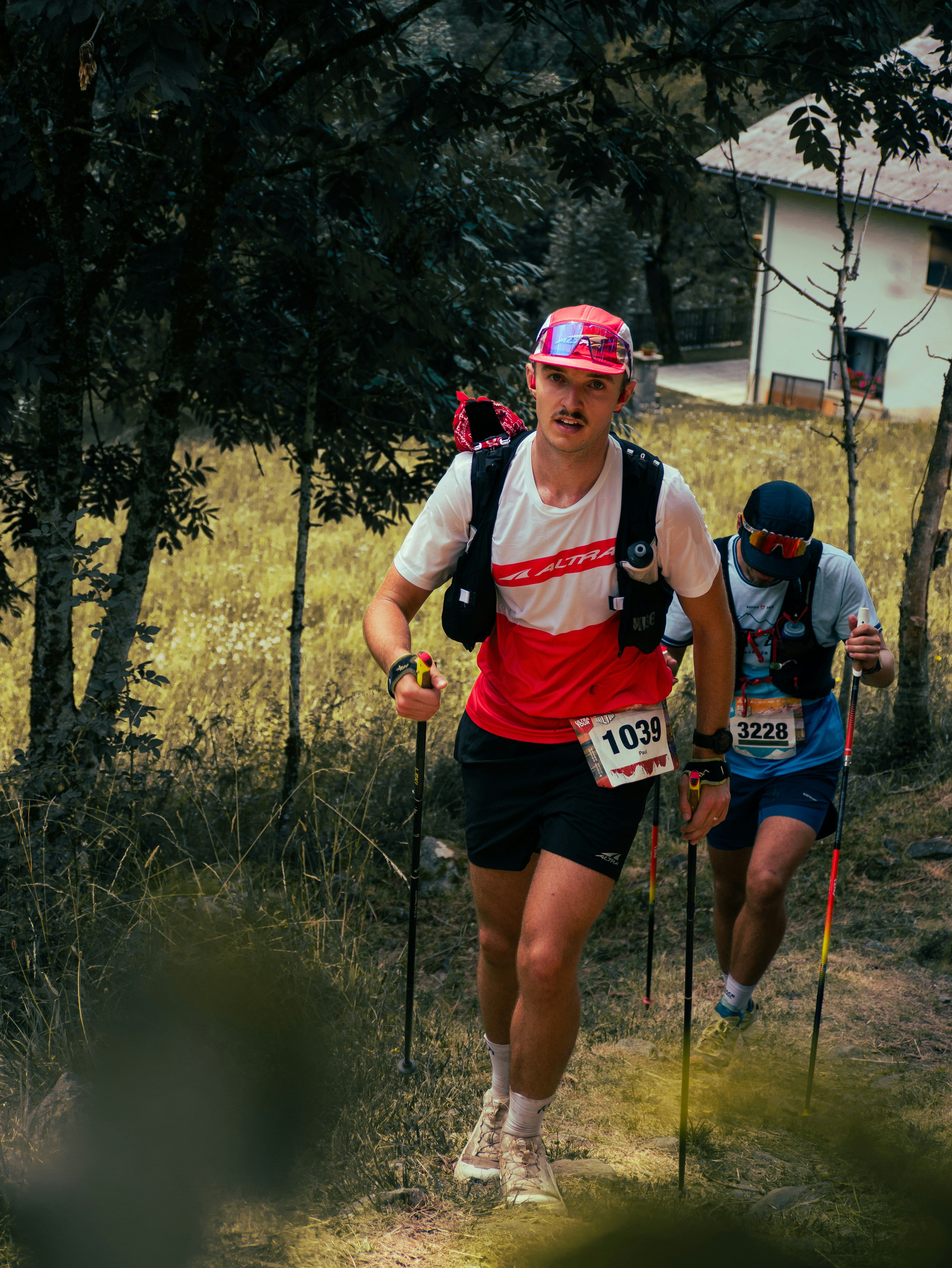 Two trail runners with trekking poles climbing a grassy hill during a race