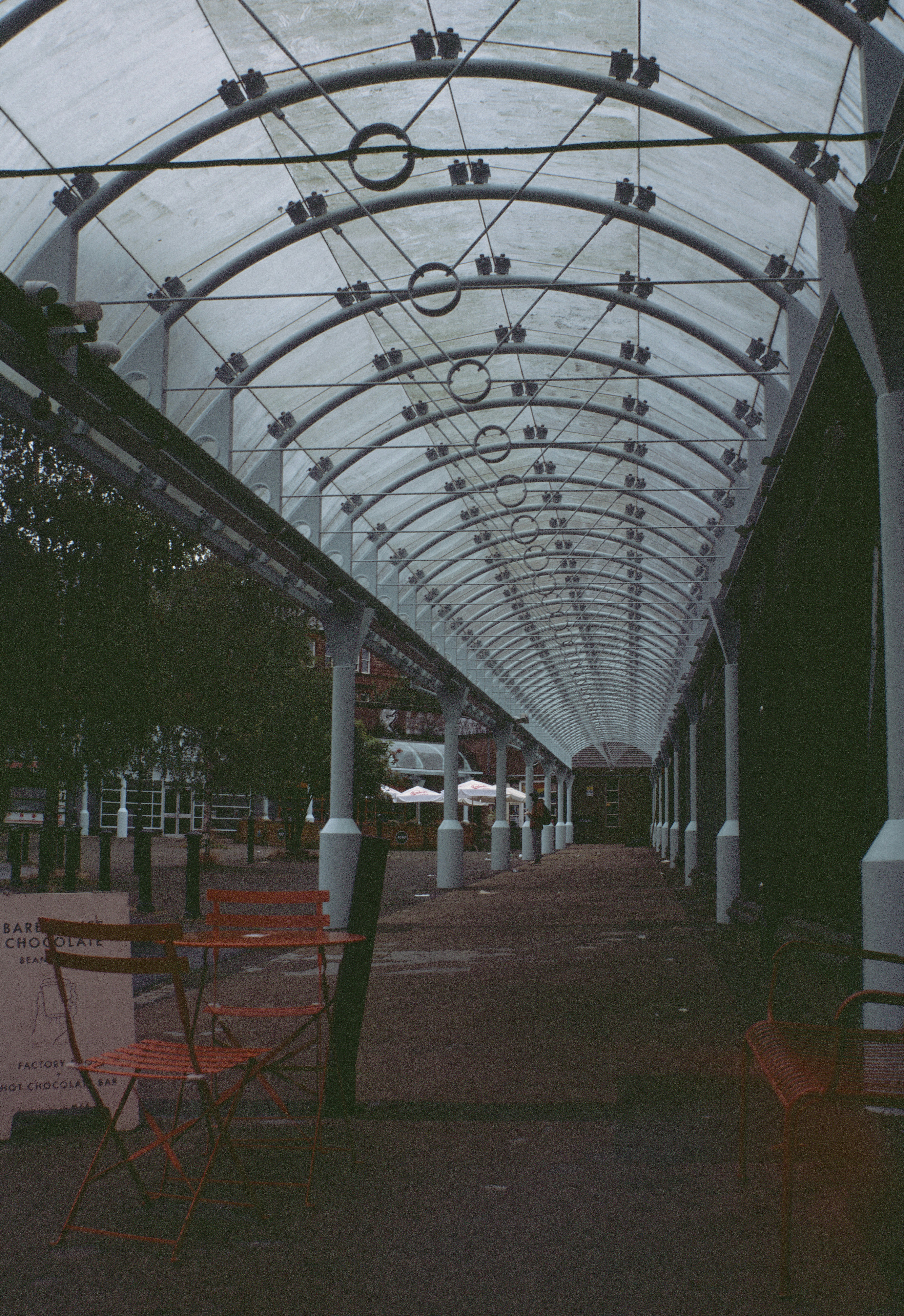 A covered walkway with a curved glass roof in Glasgow with orange chairs