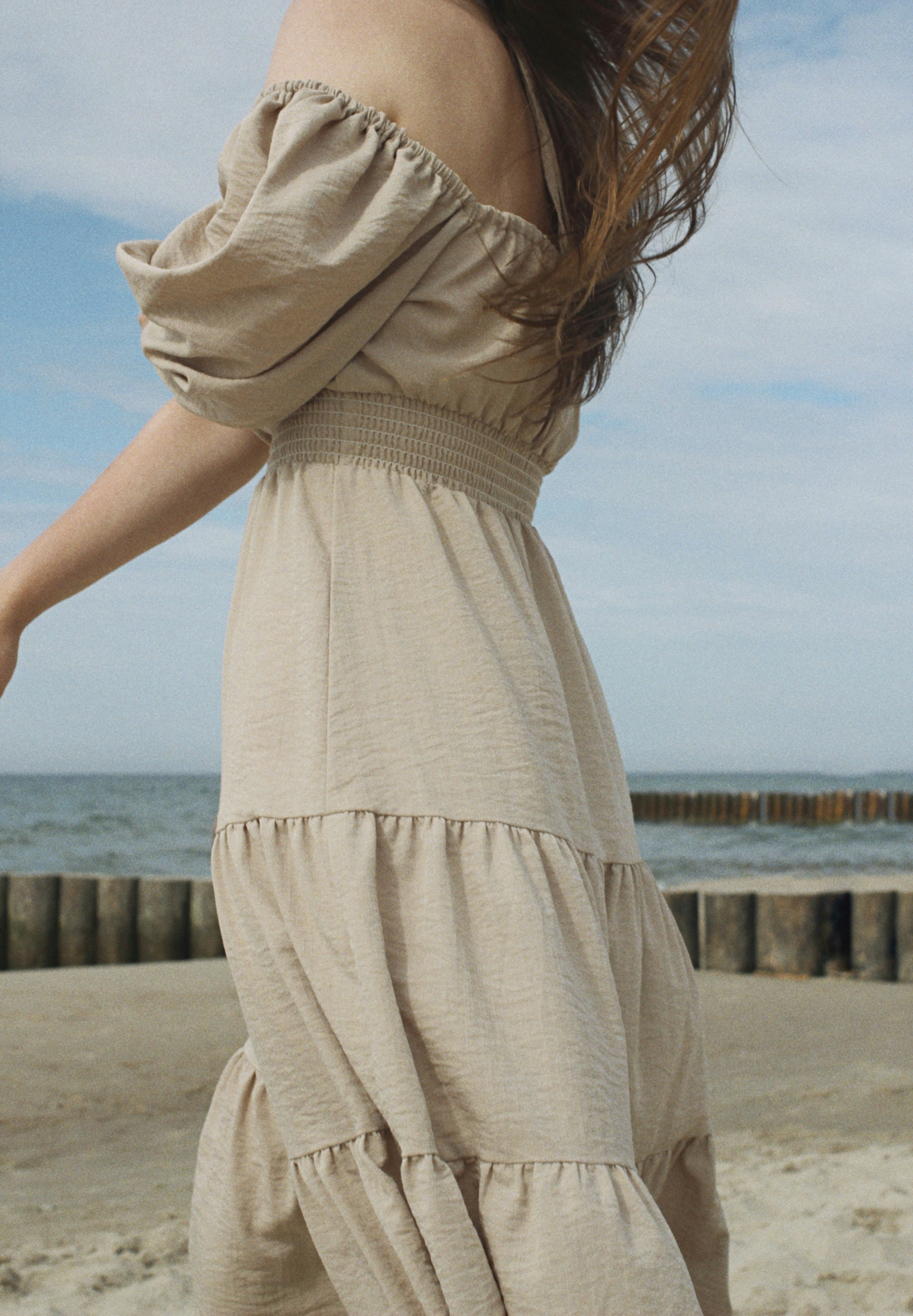 A woman in a beige off-the-shoulder dress walking on the sand