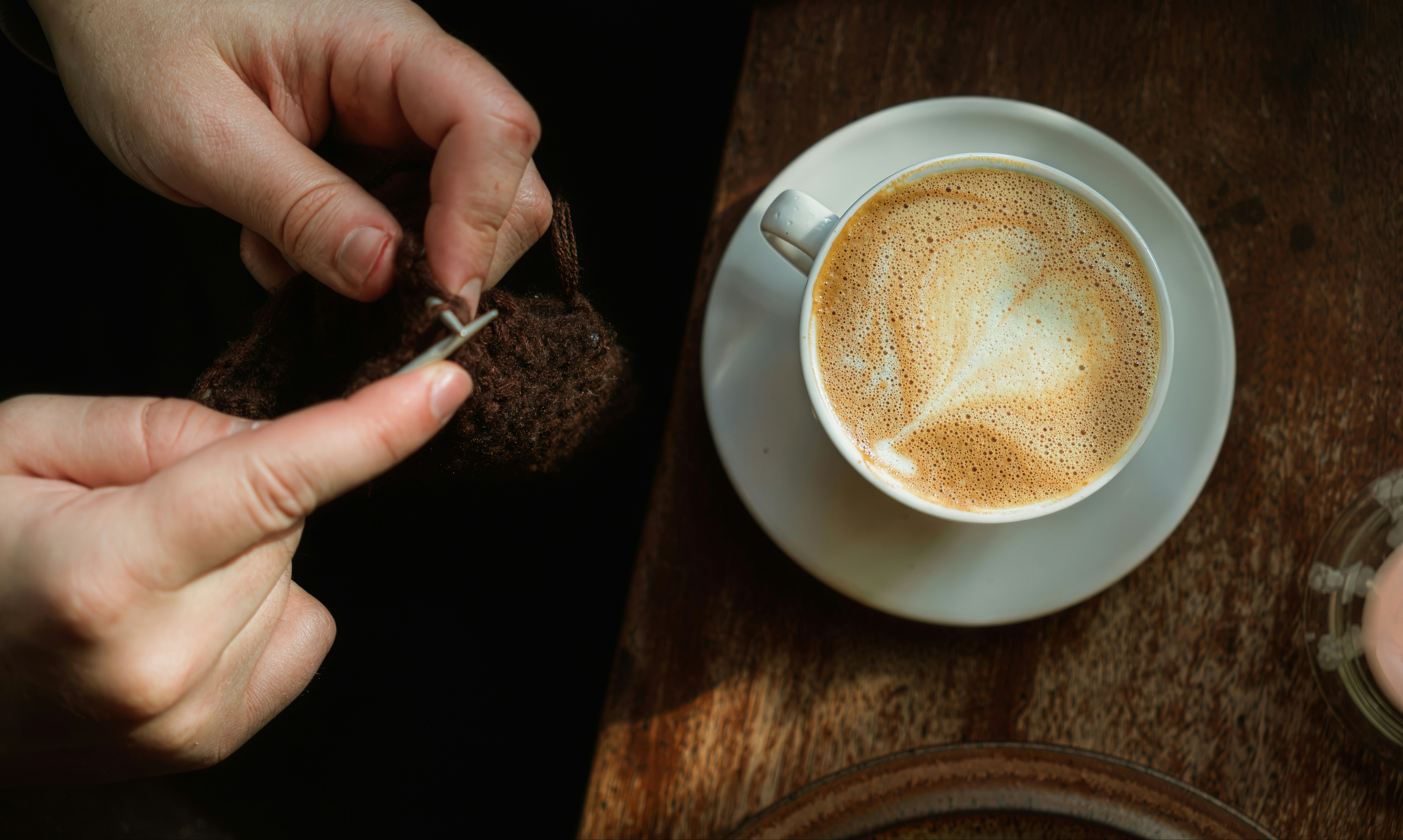Mani che lavorano a maglia con filato marrone accanto a una tazza di latte su un tavolo di legno