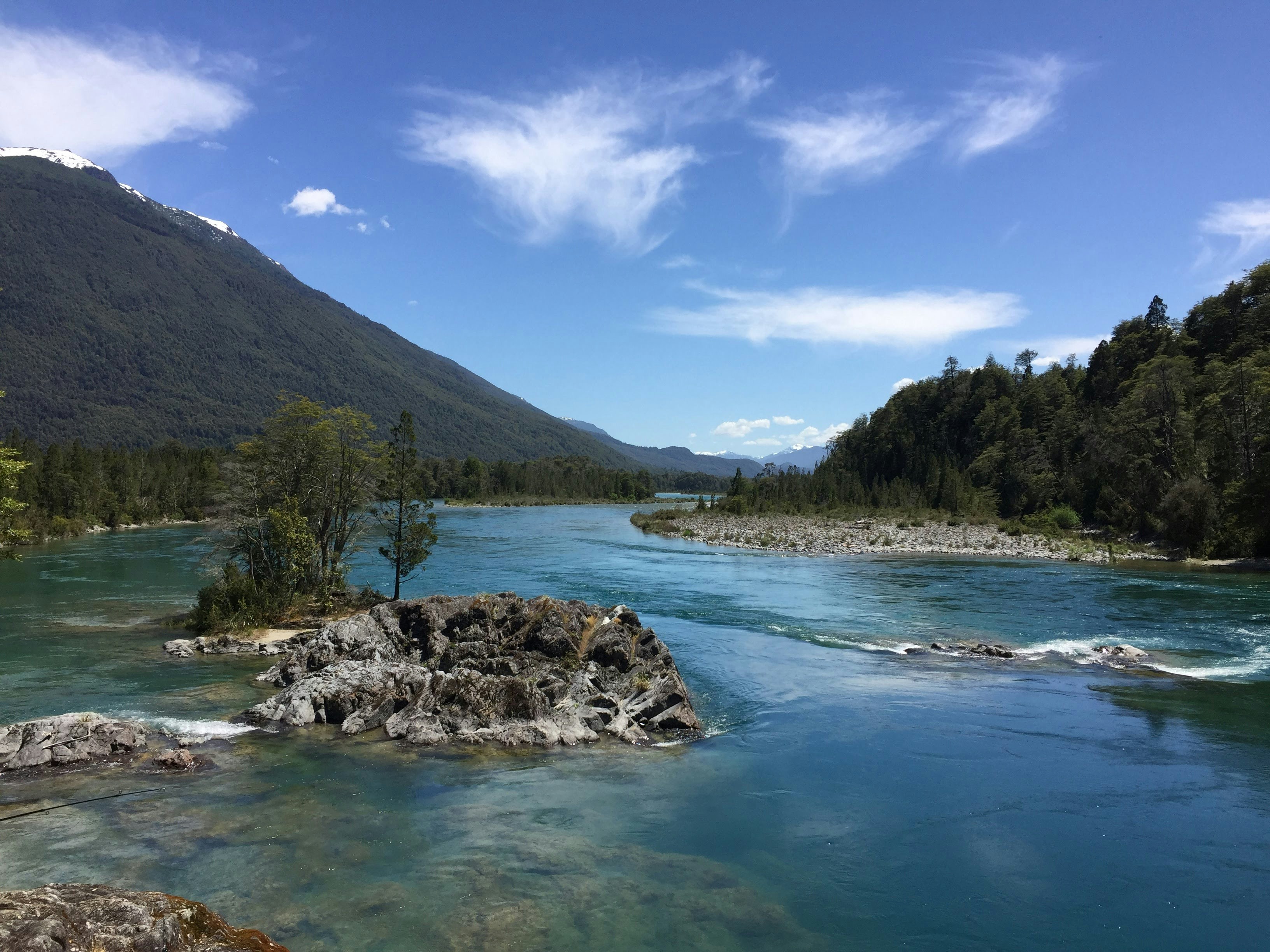 A turquoise river flowing past rocky outcrops and forested mountains under a blue sky
