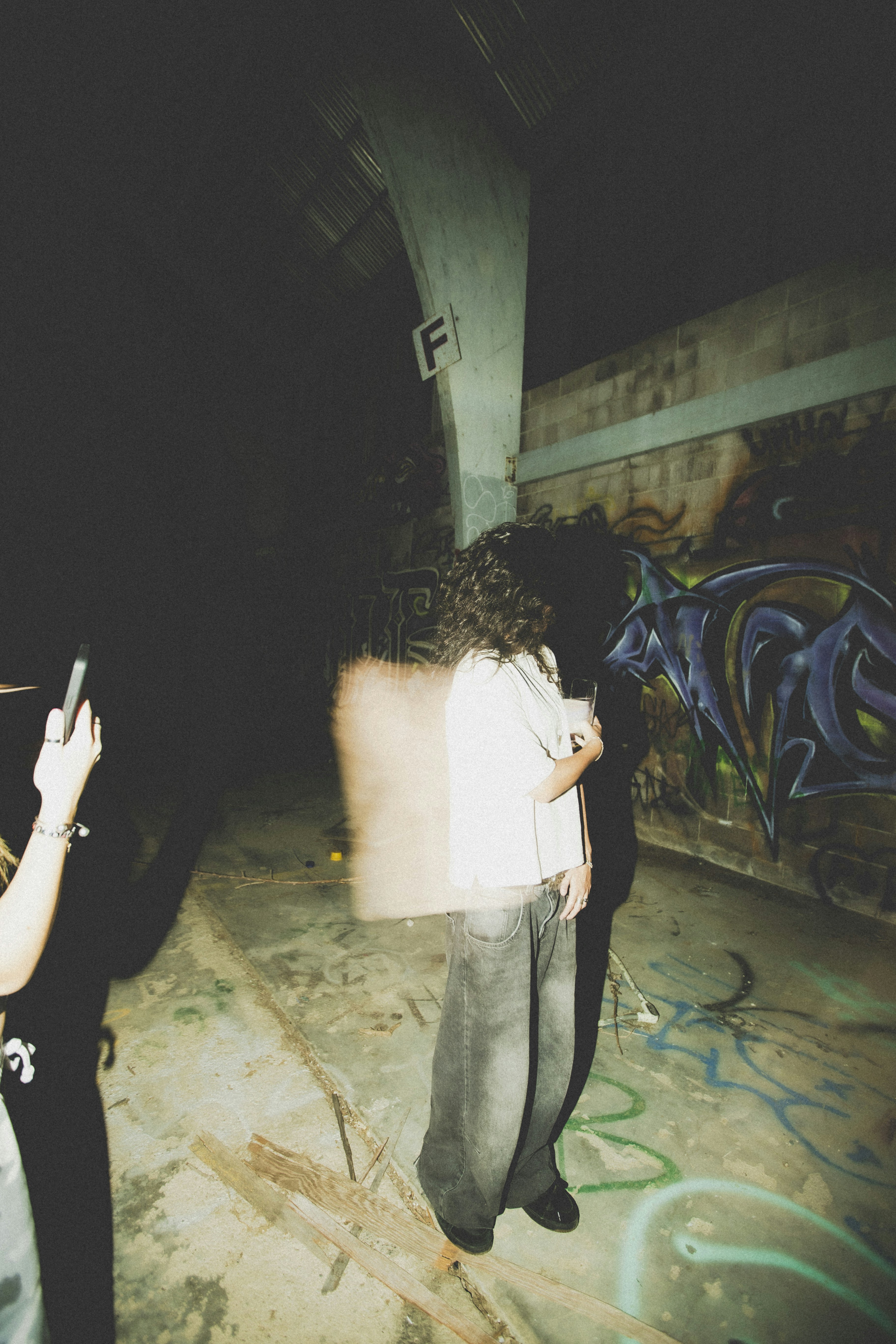 A person with long hair in a white shirt and jeans in a graffiti-covered warehouse