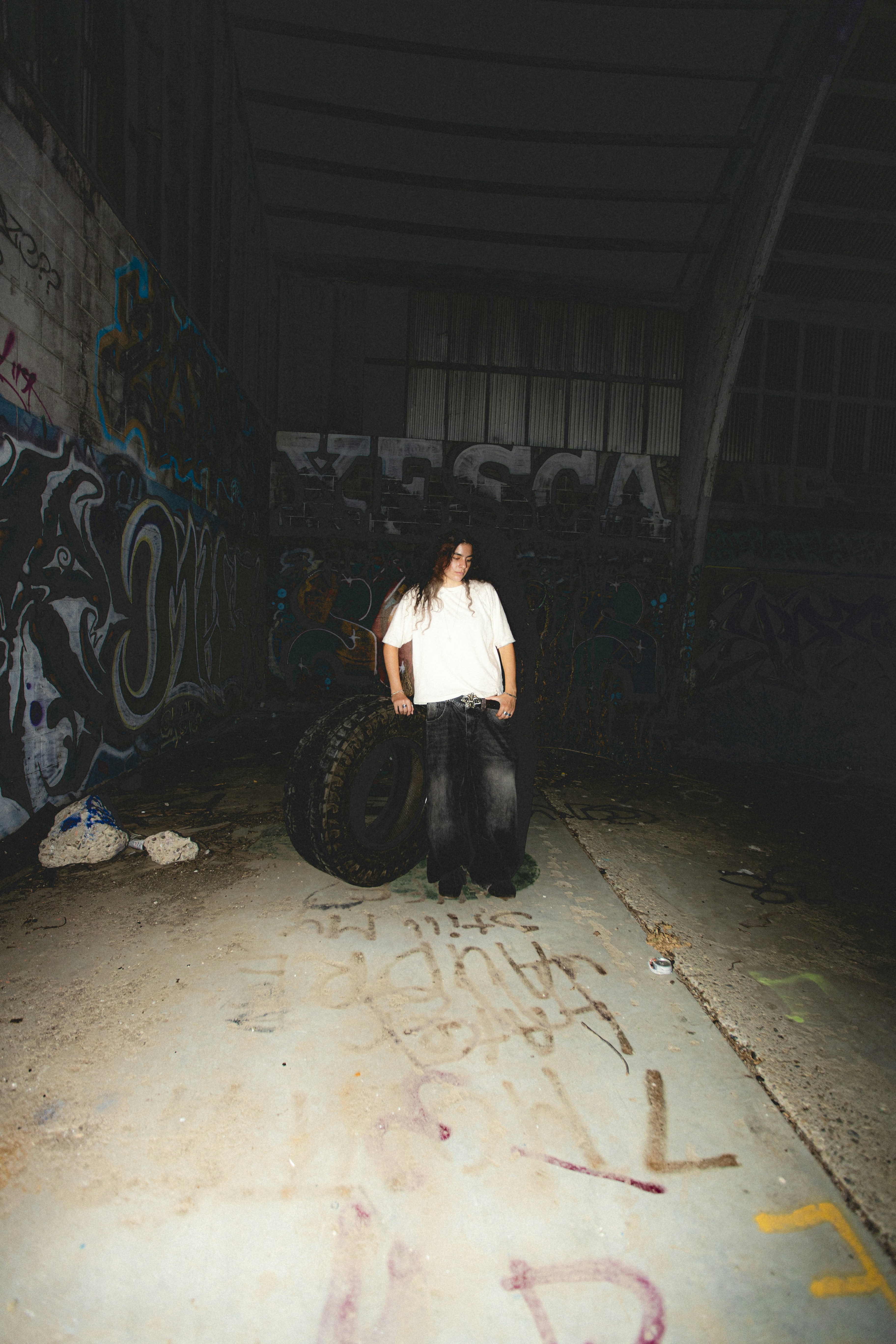 A person in a white t-shirt and dark jeans leaning on a large tire