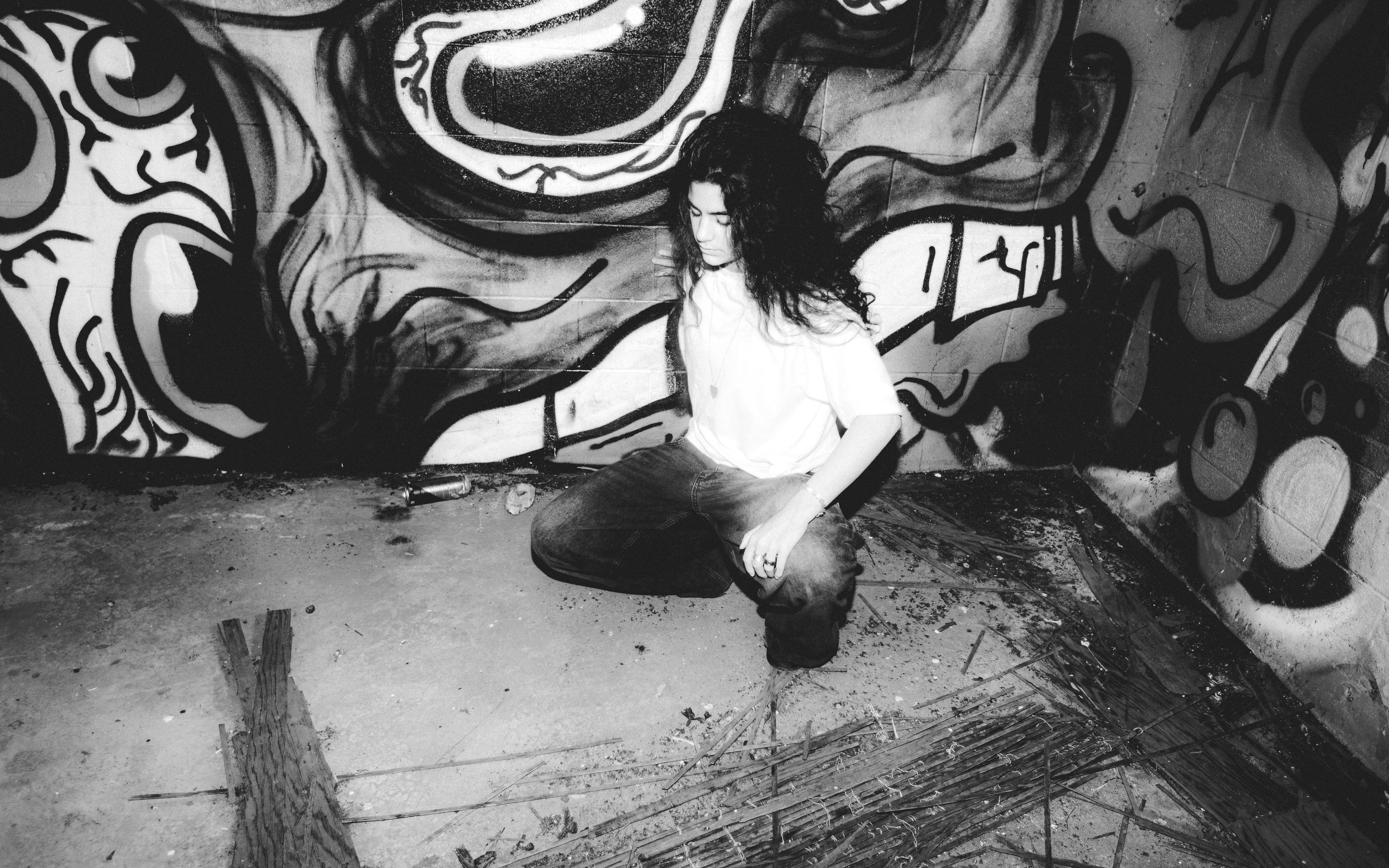 A person with long dark hair crouching before a wall covered in graffiti
