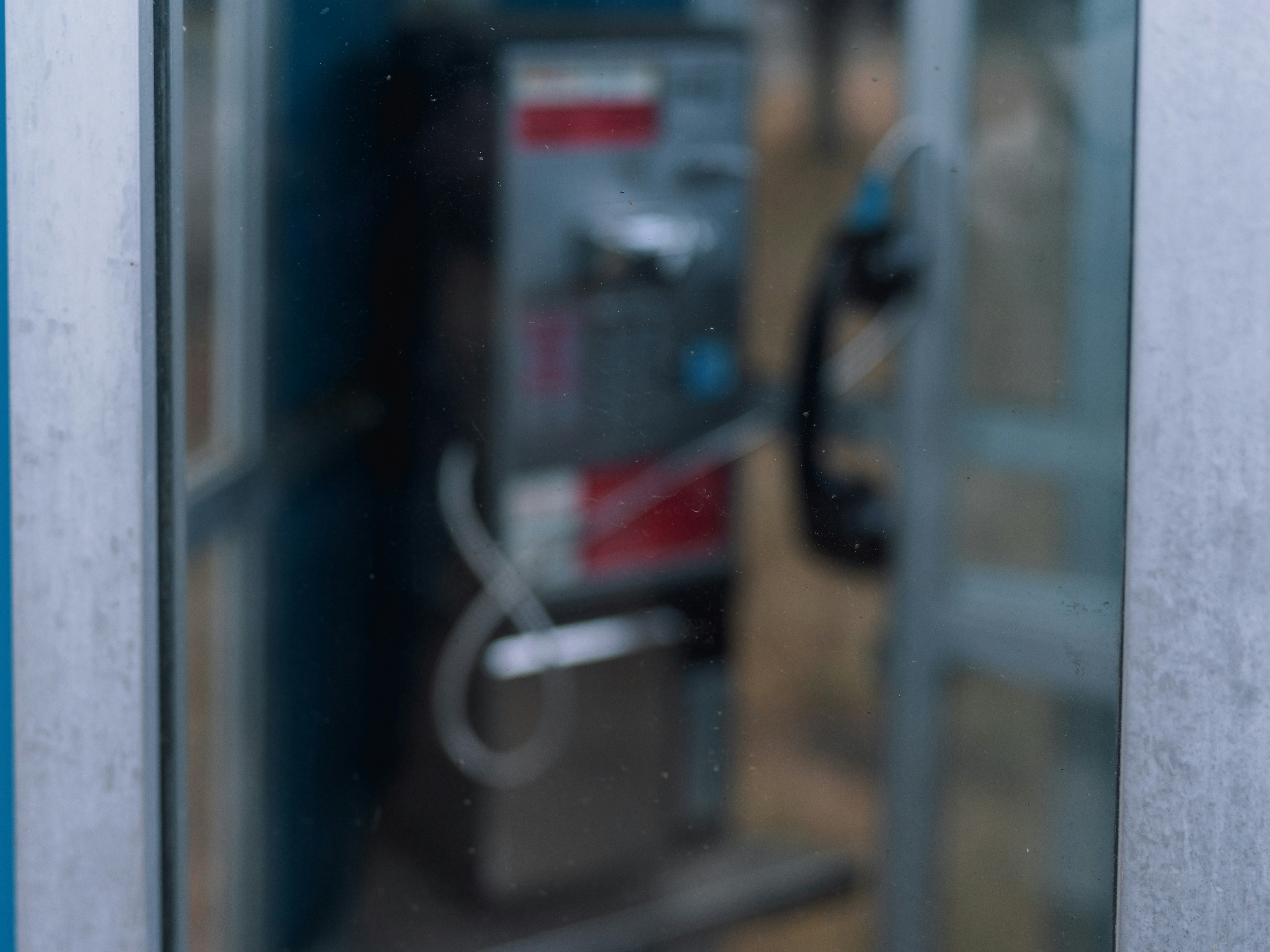 A blurred public payphone inside a glass booth with a hanging receiver