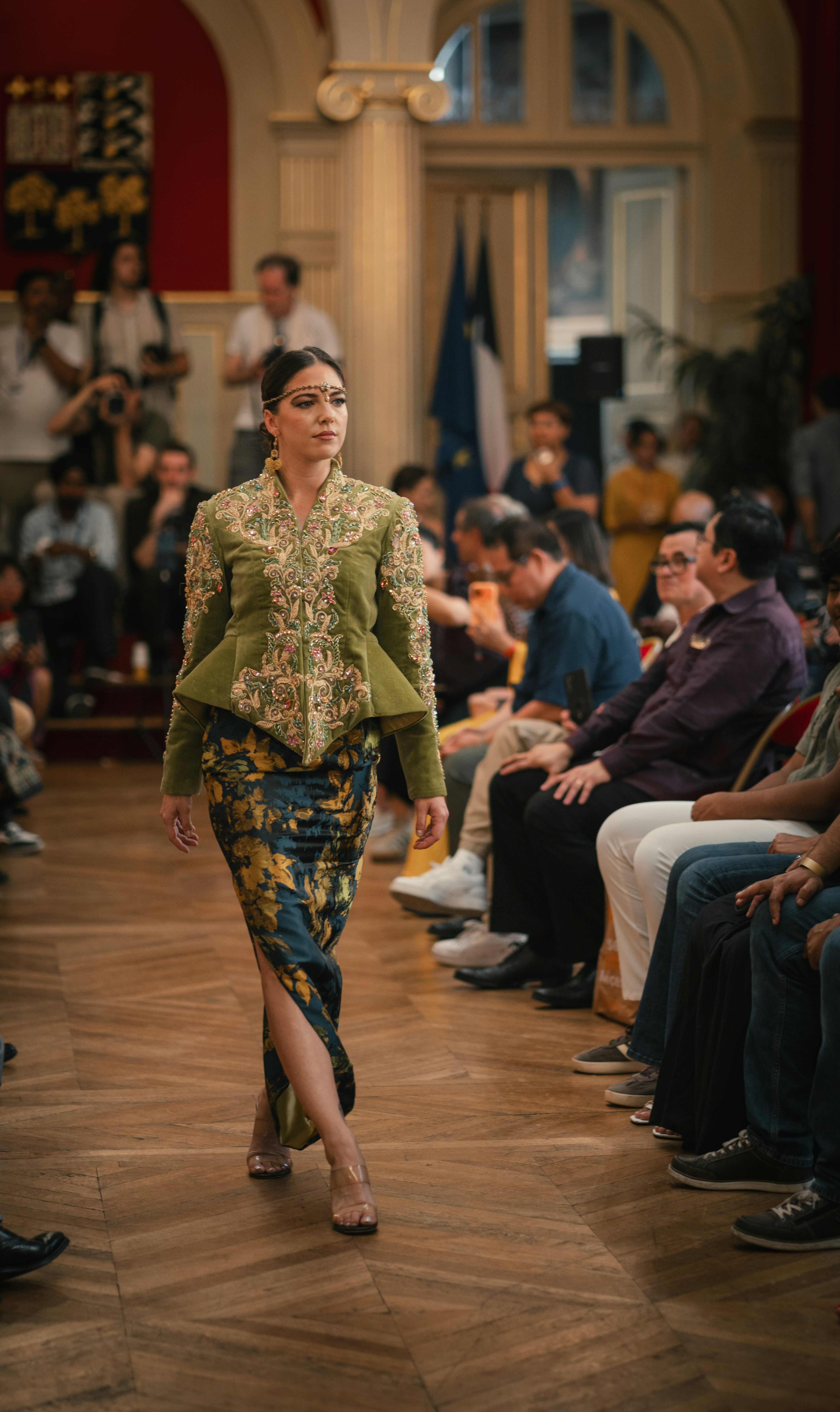 A model walking a runway in an olive green jacket and patterned skirt