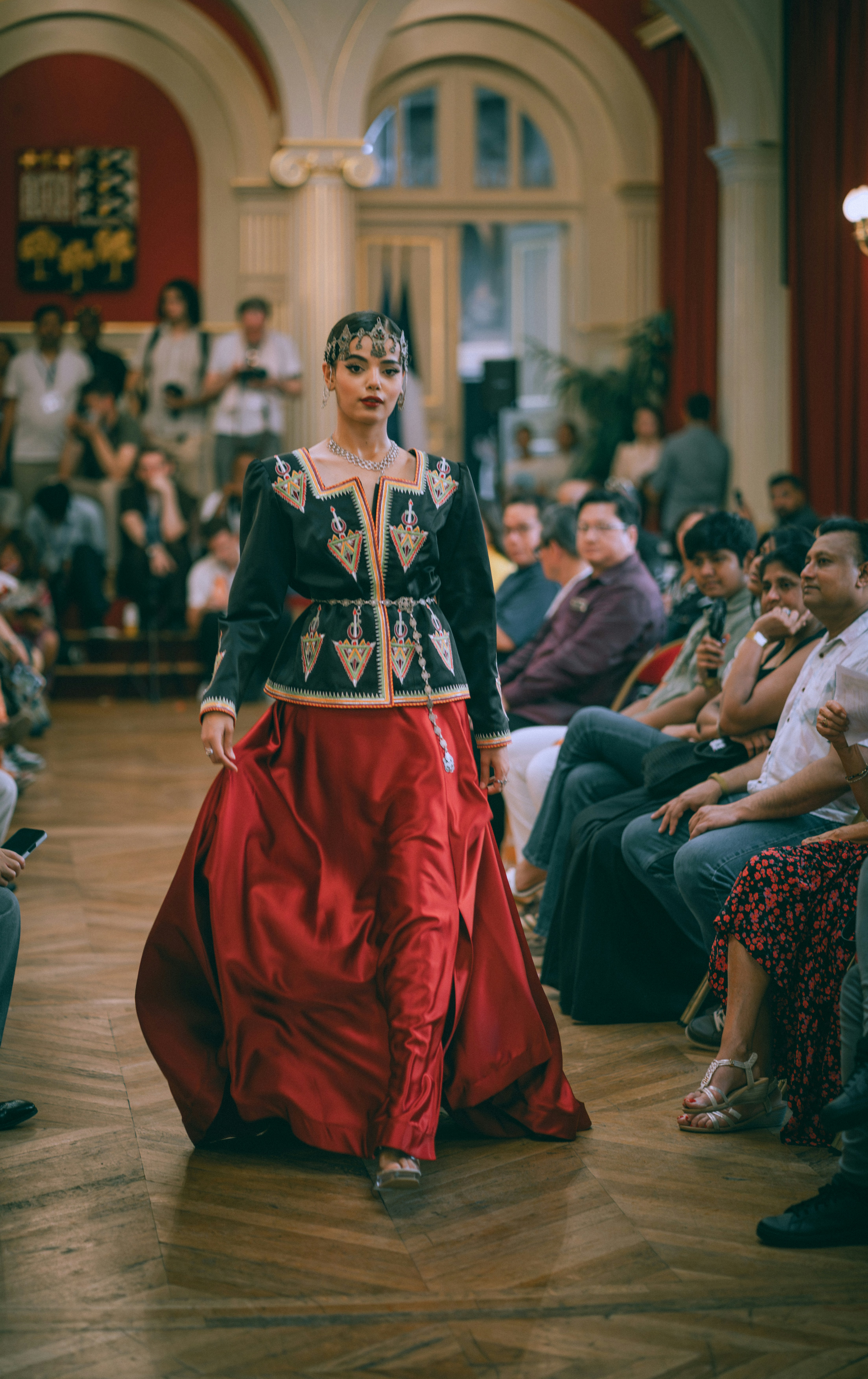 A model walking a runway in a black embroidered jacket and red skirt