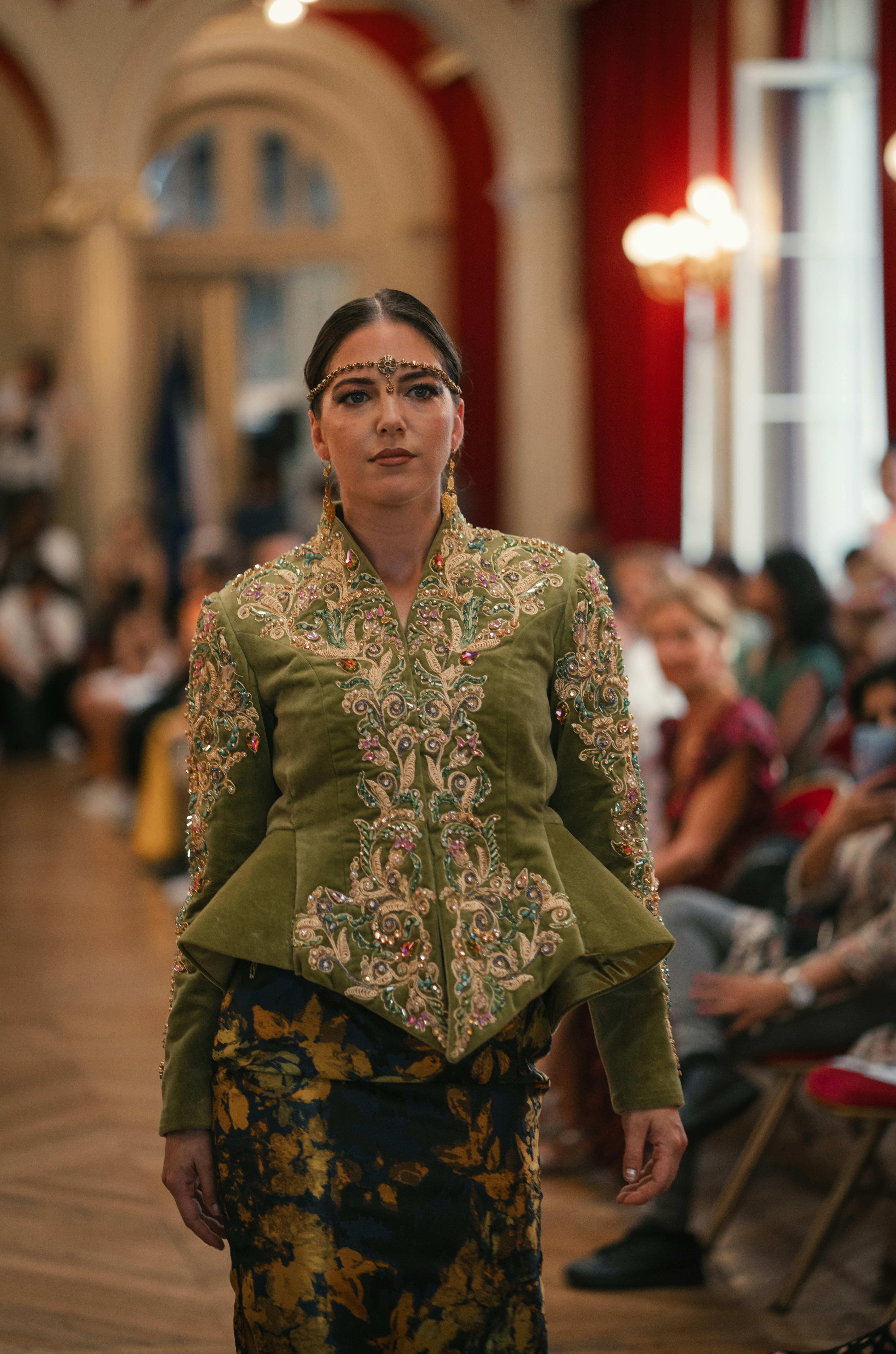 A model wearing an olive green embroidered jacket and floral skirt on a runway