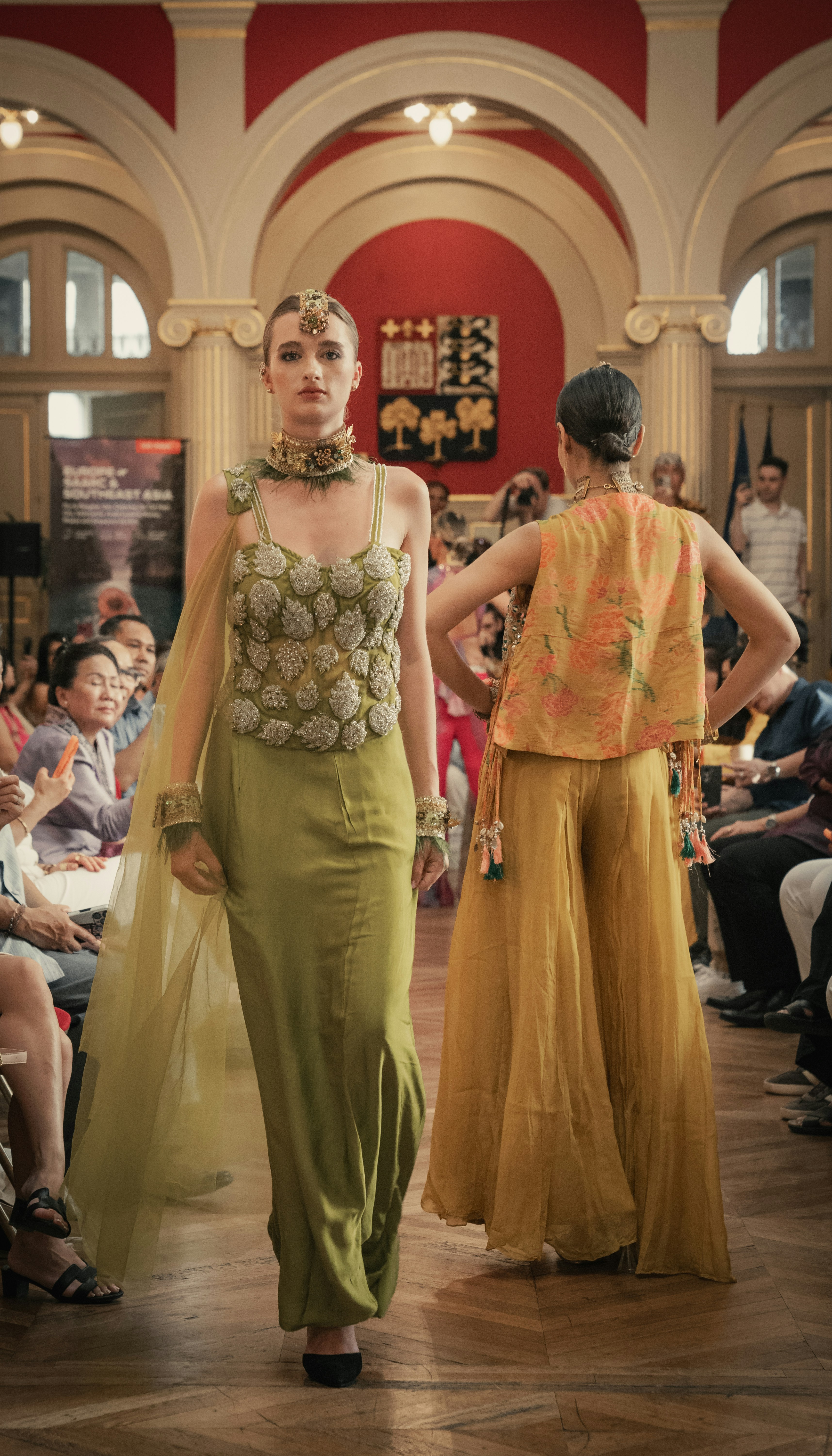 Two models walking a runway in green and yellow outfits before an audience