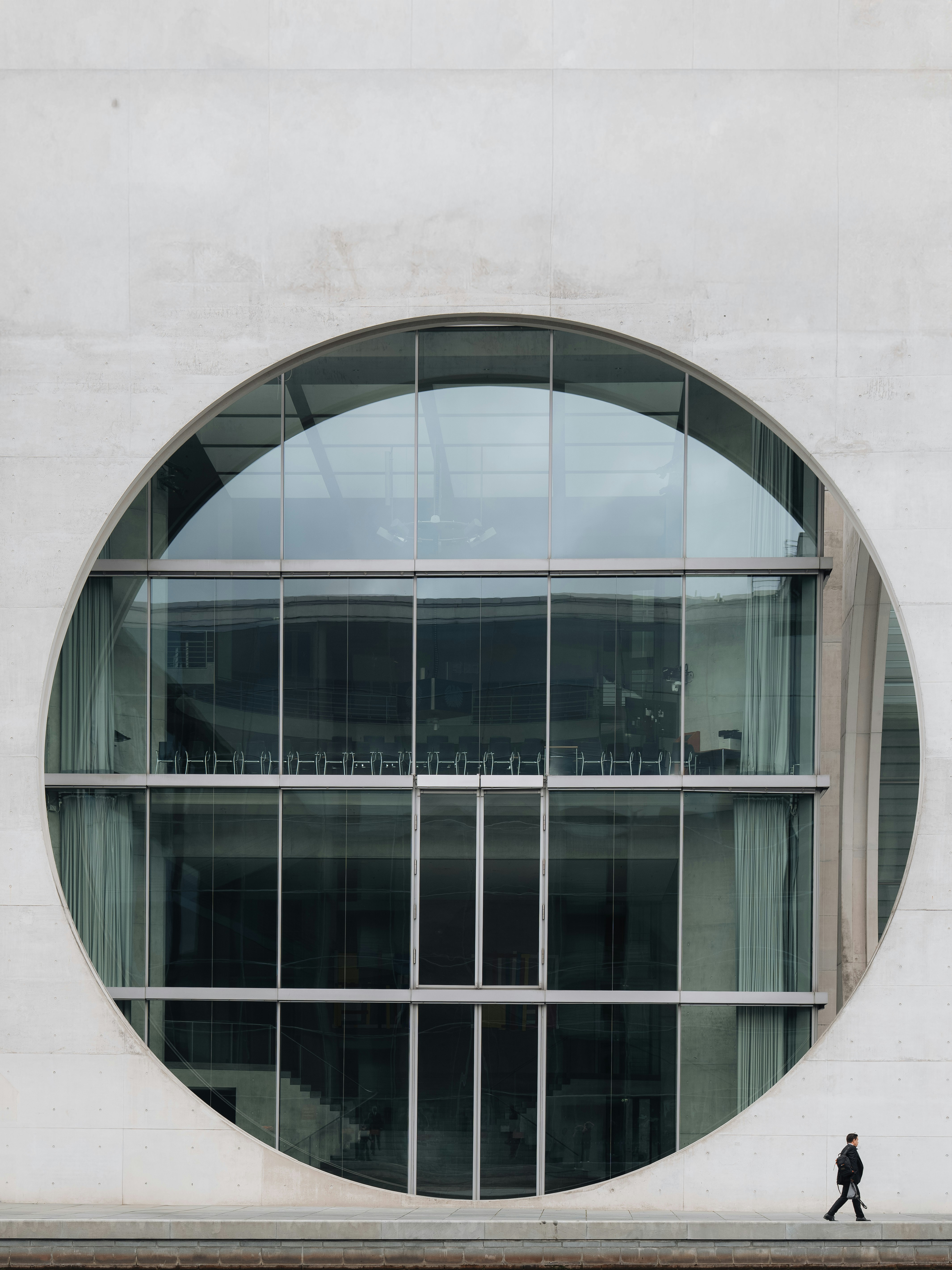 A person walking past a large circular glass window on a concrete building